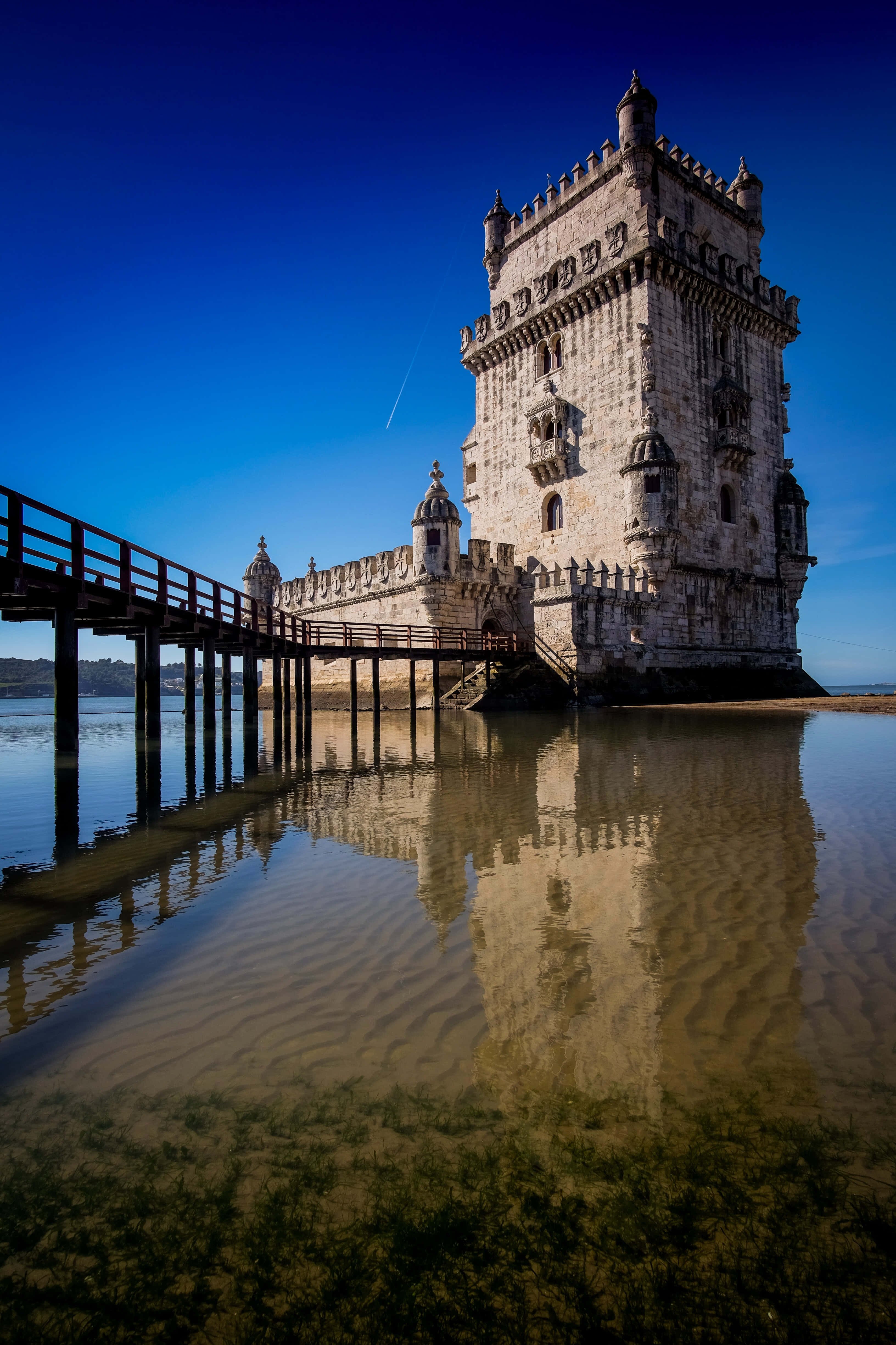 Portugal 桌布 壁紙 — 一座歷史悠久的石塔在清澈的藍天下雄偉地矗立在淺水中，其倒影在前方泛起漣漪。