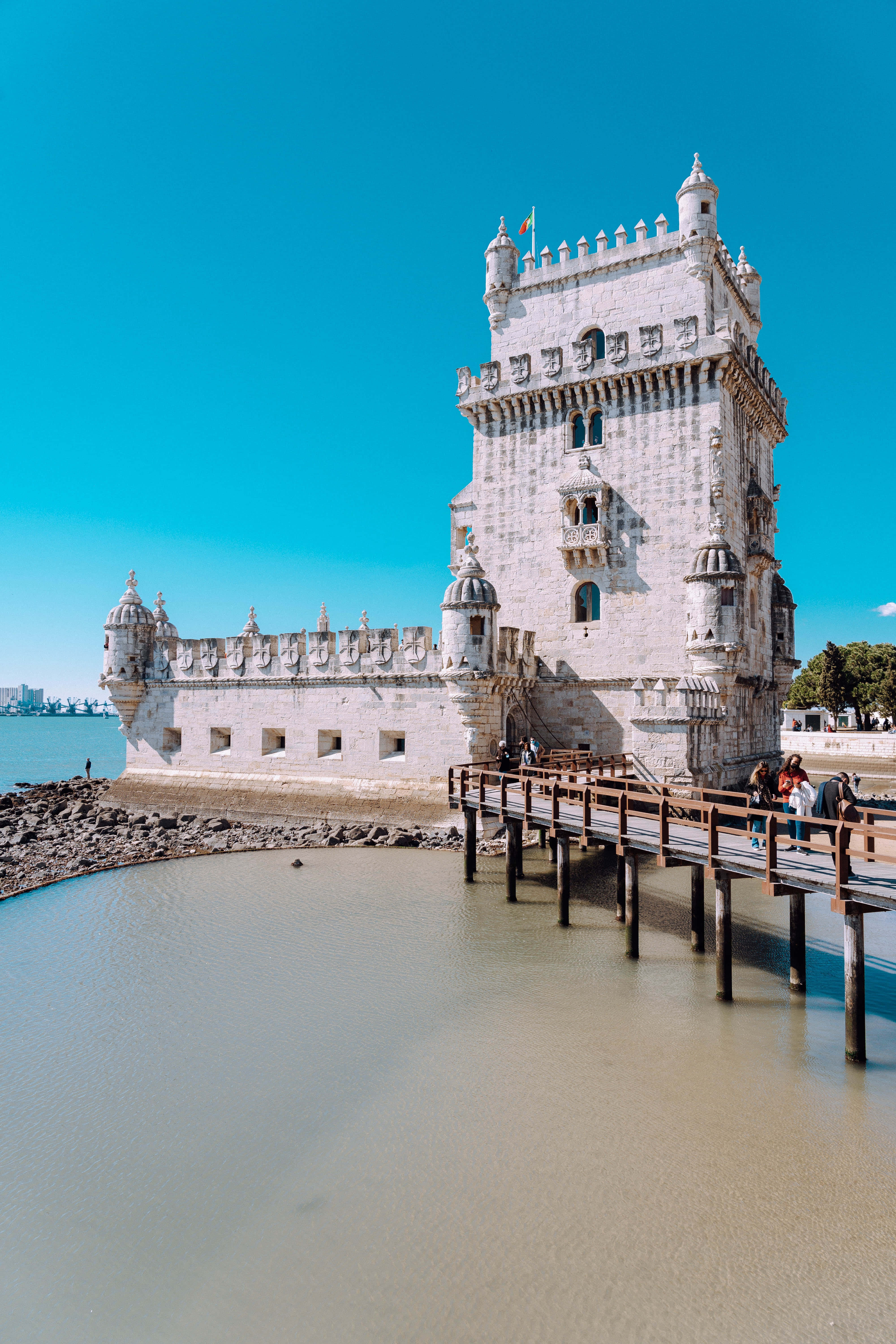 Portugal 桌布 壁紙 — 一座歷史悠久的石塔在清澈藍天下雄偉地矗立在水邊，遊客們正走過木製碼頭。