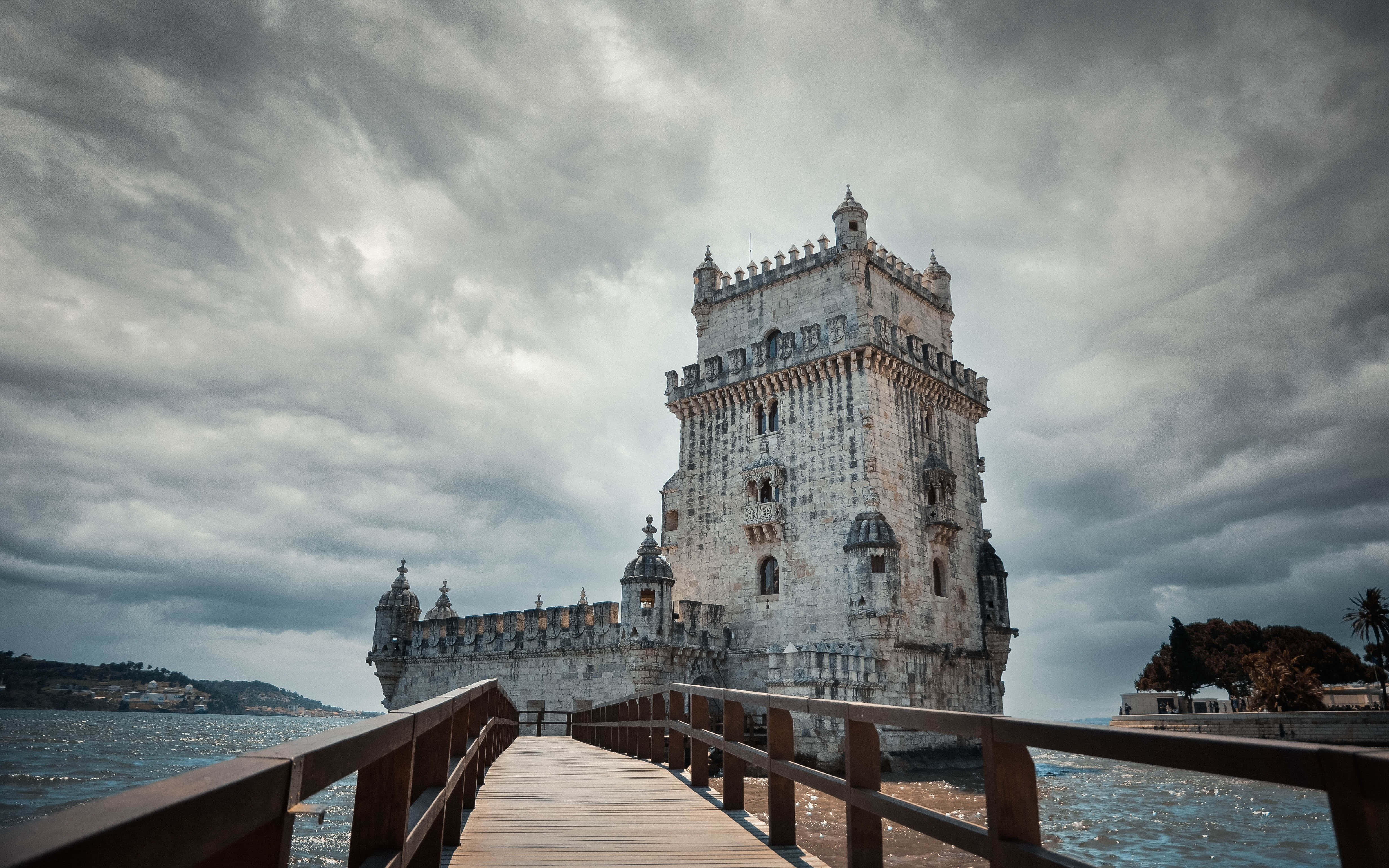Portugal 桌布 壁紙 — 一座歷史悠久的石塔在陰沉灰暗的天空下莊嚴矗立，可經由延伸至平靜水面的木製碼頭進入。