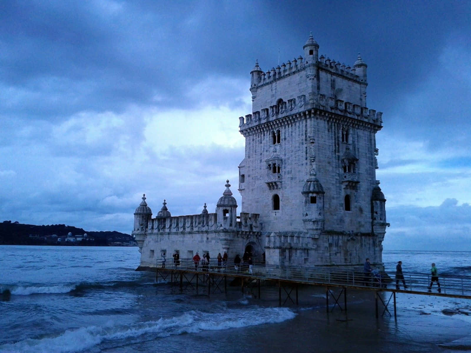 Portugal 桌布 壁紙 — 一座歷史悠久的石塔孤立在水中，背景是陰沉的暮色天空，人們沿著木碼頭行走。