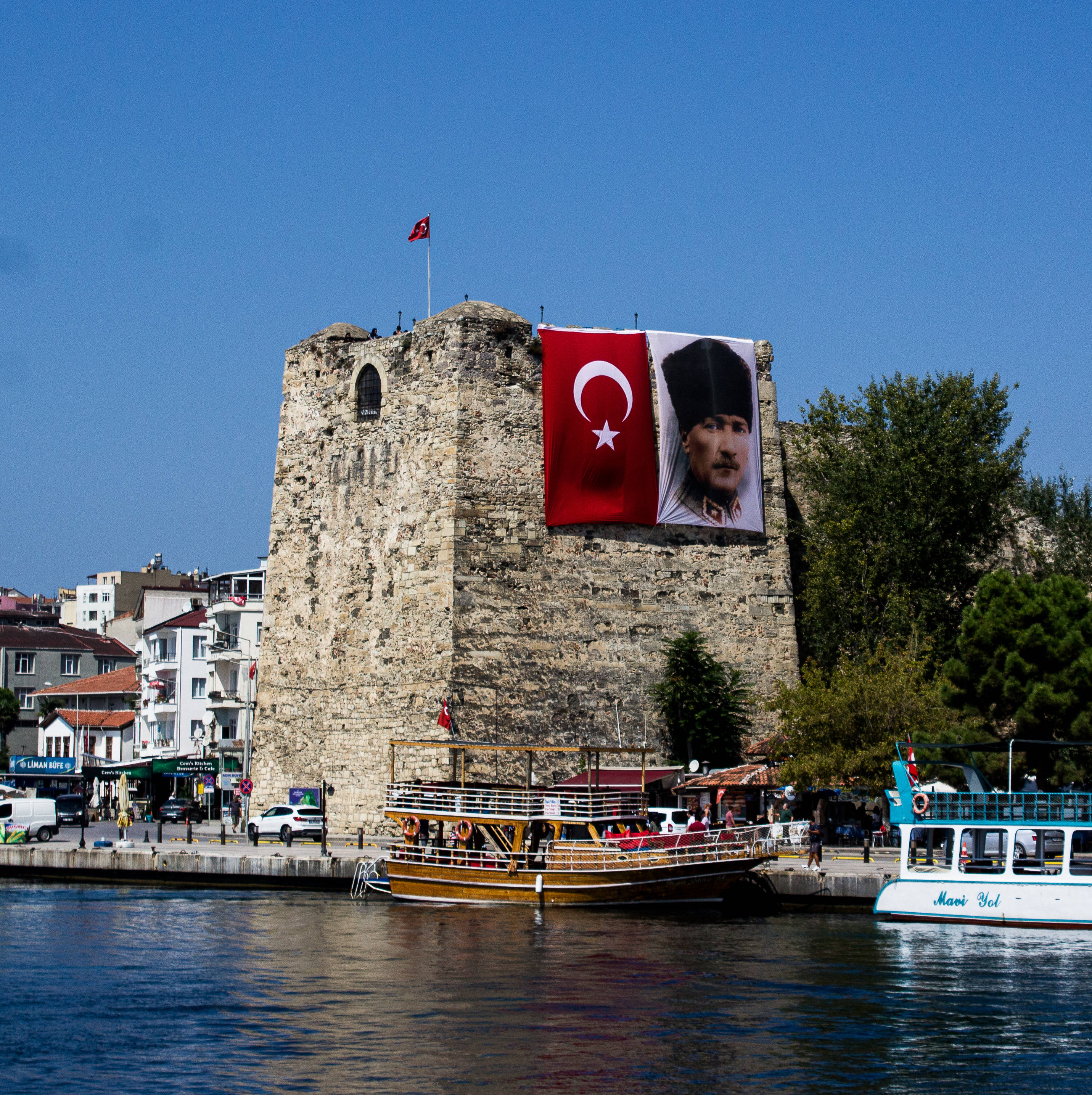 Ataturk 桌布 壁紙 — 一座歷史悠久的石塔矗立在水邊，展示著一面巨大的土耳其國旗和凱末爾肖像，在晴朗的藍天下，附近停泊著船隻。