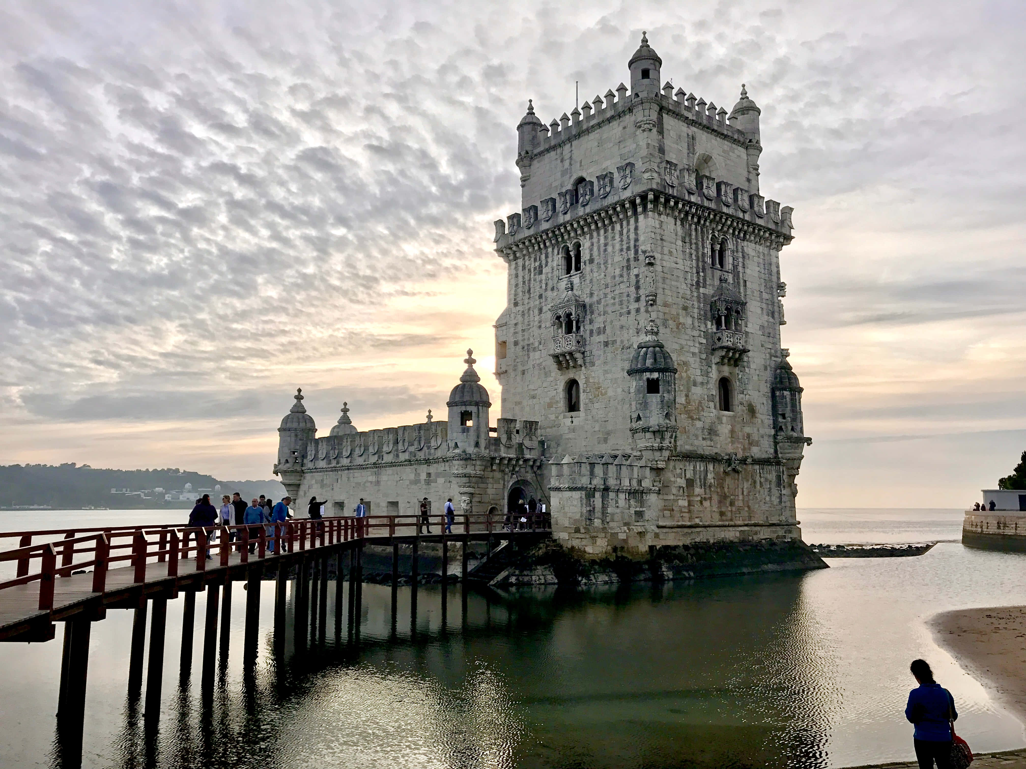 Portugal 桌布 壁紙 — 一座歷史悠久的石塔矗立在水面上，由木製碼頭連接，天空多雲，暮色蒼茫。