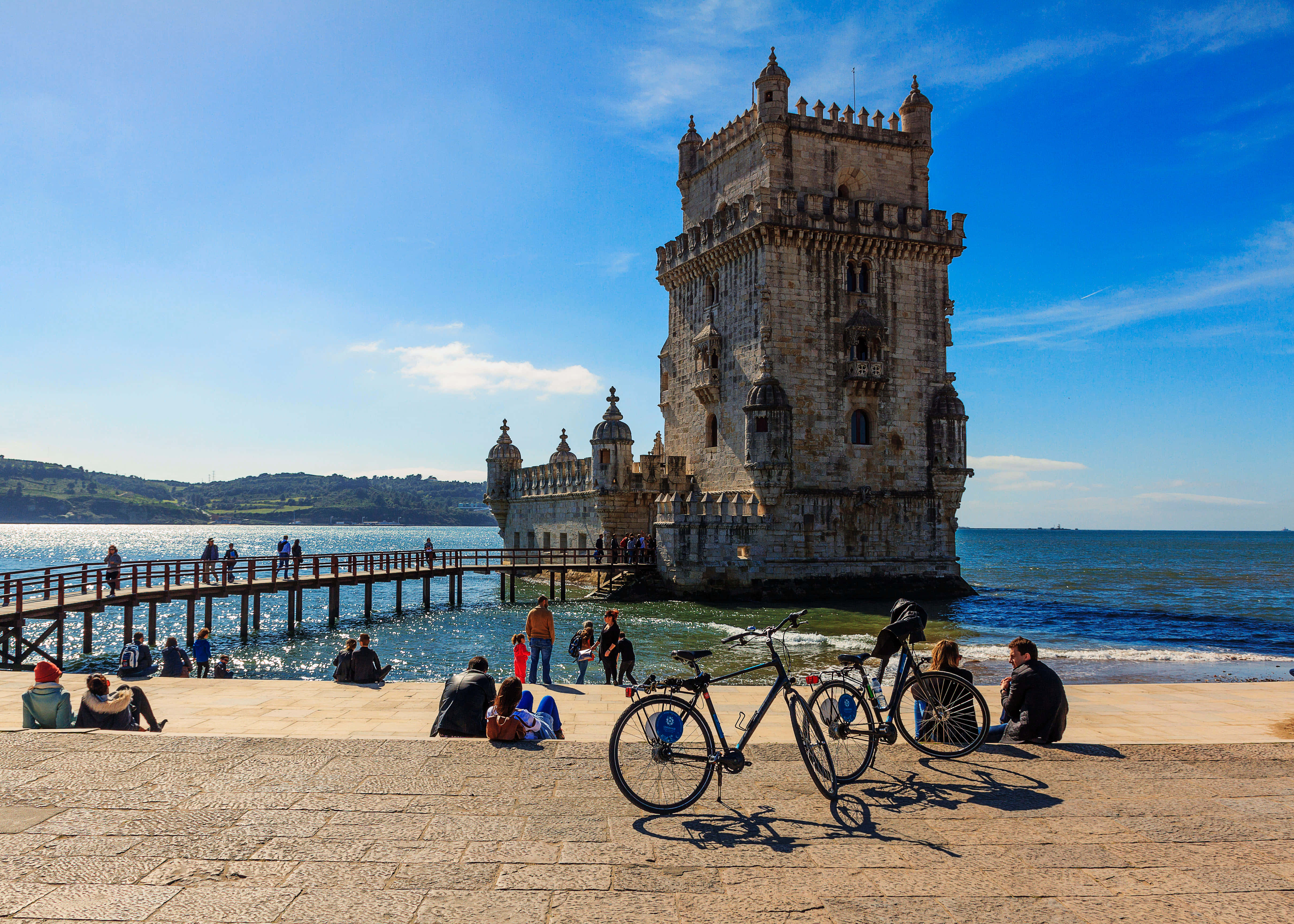 Portugal 桌布 壁紙 — 一座歷史悠久的石塔矗立在海中，由木製碼頭連接，人們在晴朗的藍天下在石頭長廊上放鬆。