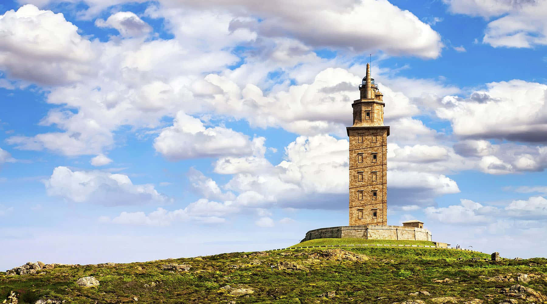 Lighthouse 桌布 壁紙 — 一座歷史悠久的石塔矗立在長滿青草的山丘上，天空湛藍，點綴著蓬鬆的白雲。