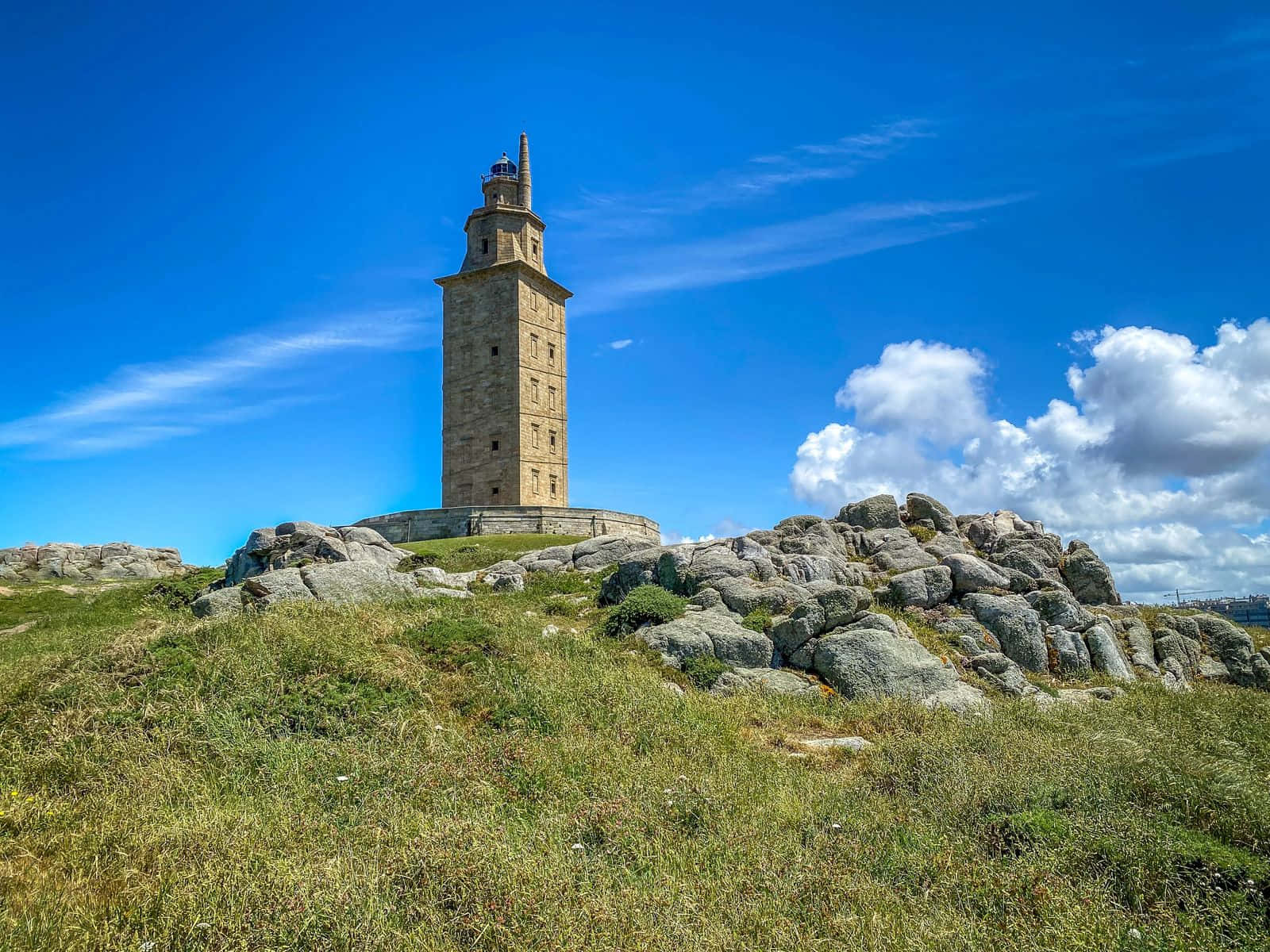 Lighthouse 桌布 壁紙 — 一座歷史悠久的石塔聳立在岩石山上，天空湛藍，白雲朵朵。
