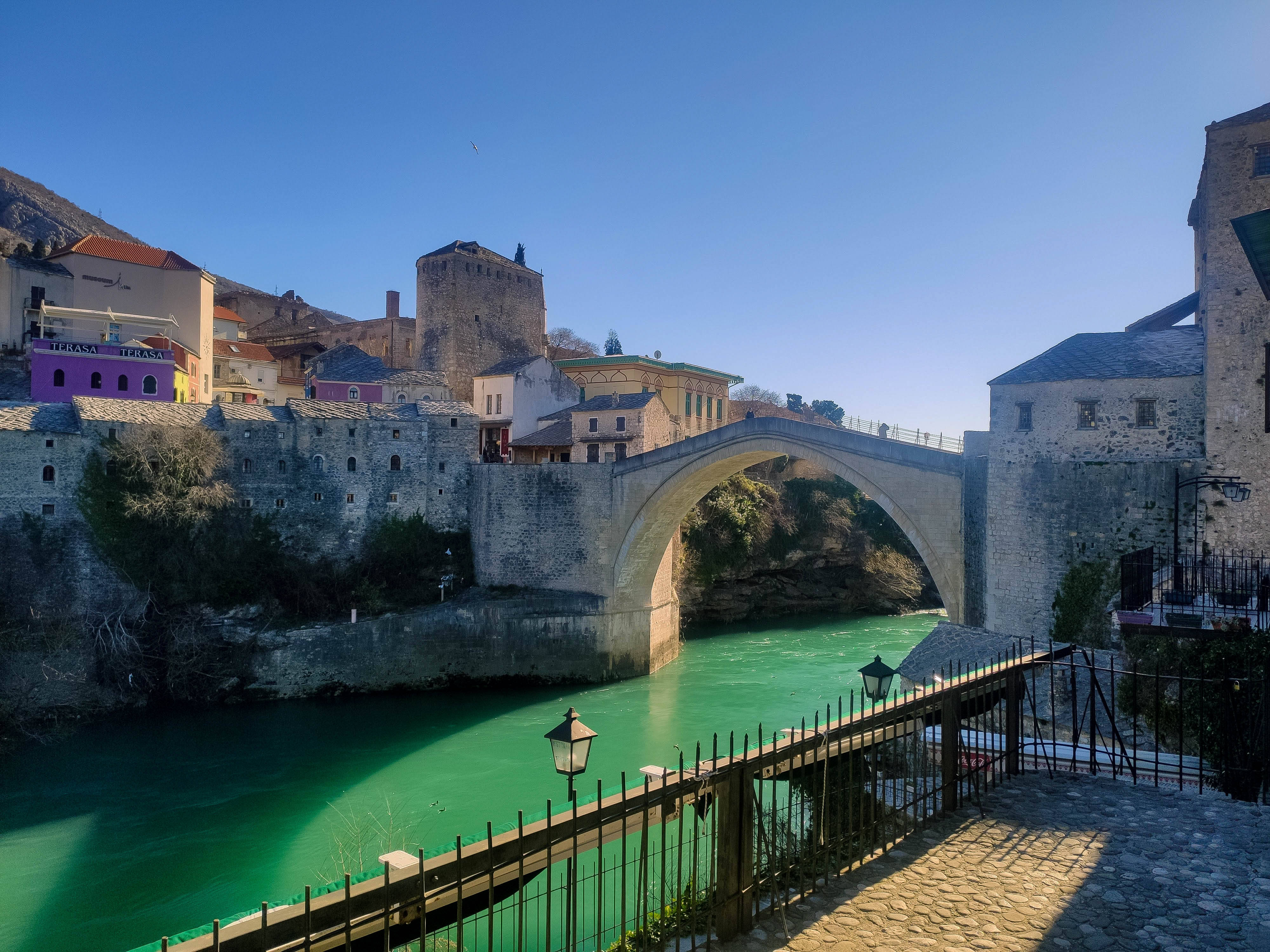 Bosnia And Herzegovina 桌布 壁紙 — 一座歷史悠久的石橋拱跨在充滿活力的碧綠河流上，天空清澈湛藍，河岸兩旁排列著古老的建築。