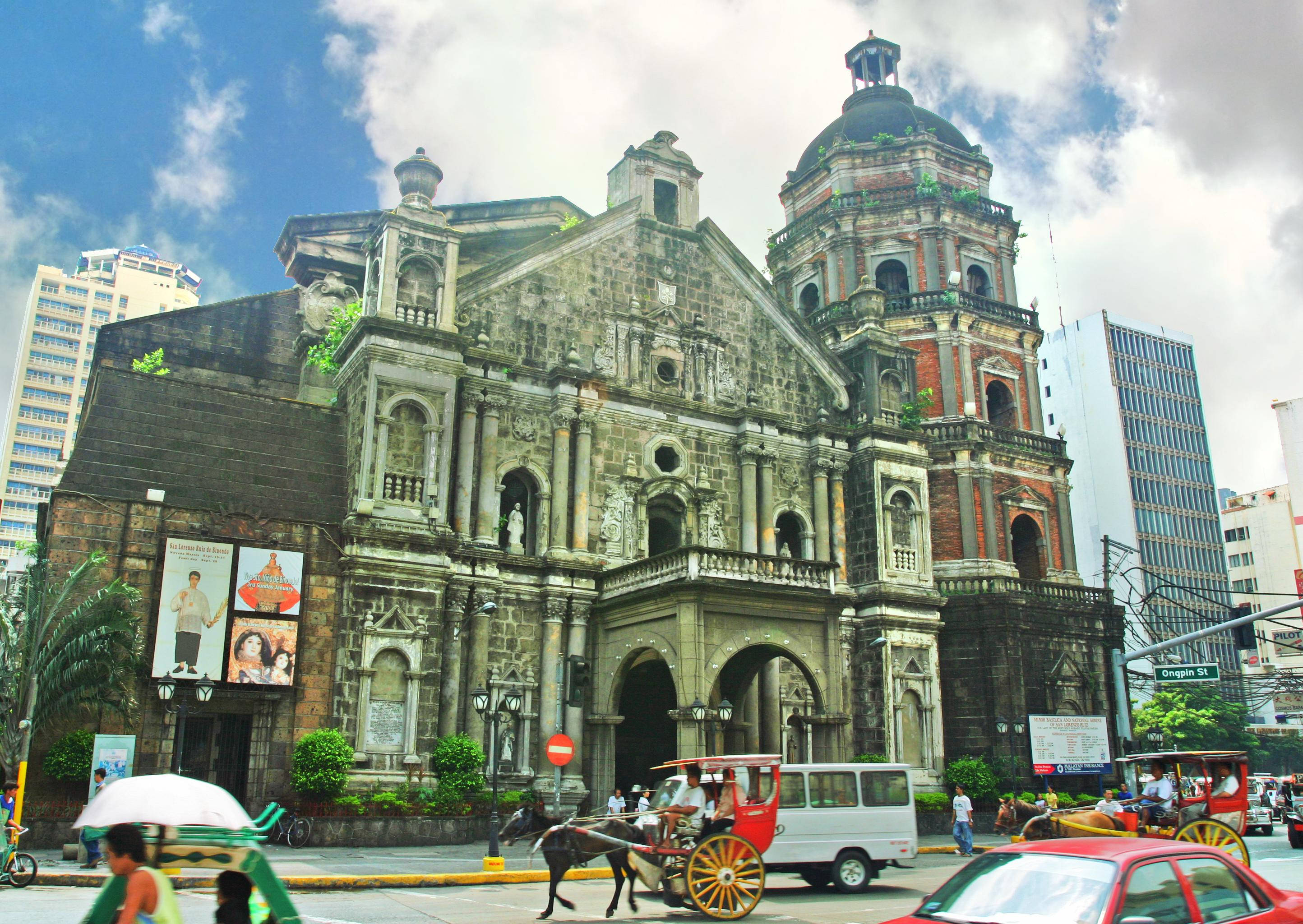 Manila 桌布 壁紙 — 一座歷史悠久的石砌教堂，帶有圓頂塔樓，聳立在繁華的城市街道上，馬車和現代汽車在部分多雲的天空下穿行。