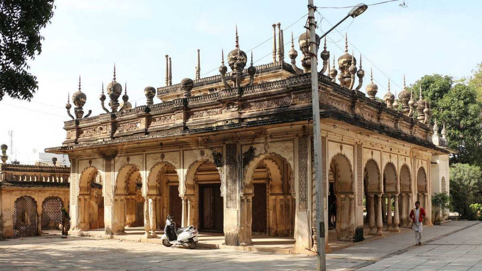 Hyderabad 桌布 壁紙 — 一座歷史悠久的石造清真寺，擁有拱形柱廊和華麗的圓頂屋頂，在明亮的日光下矗立，附近停著一輛摩托車。