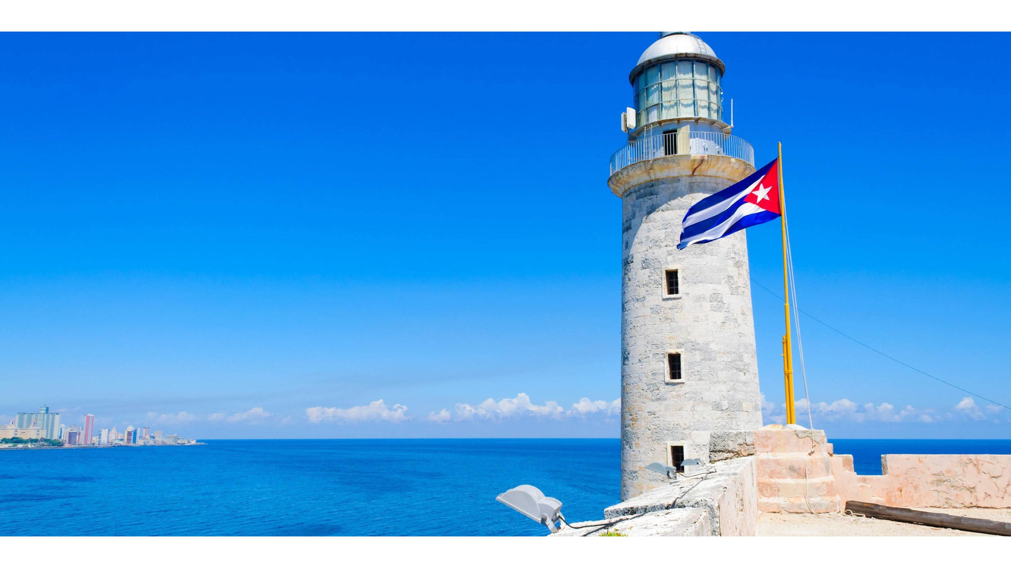 Cuba 桌布 壁紙 — 一座歷史悠久的石造燈塔矗立在海岸懸崖上，古巴國旗在清澈的藍天和碧綠的海洋中飄揚。