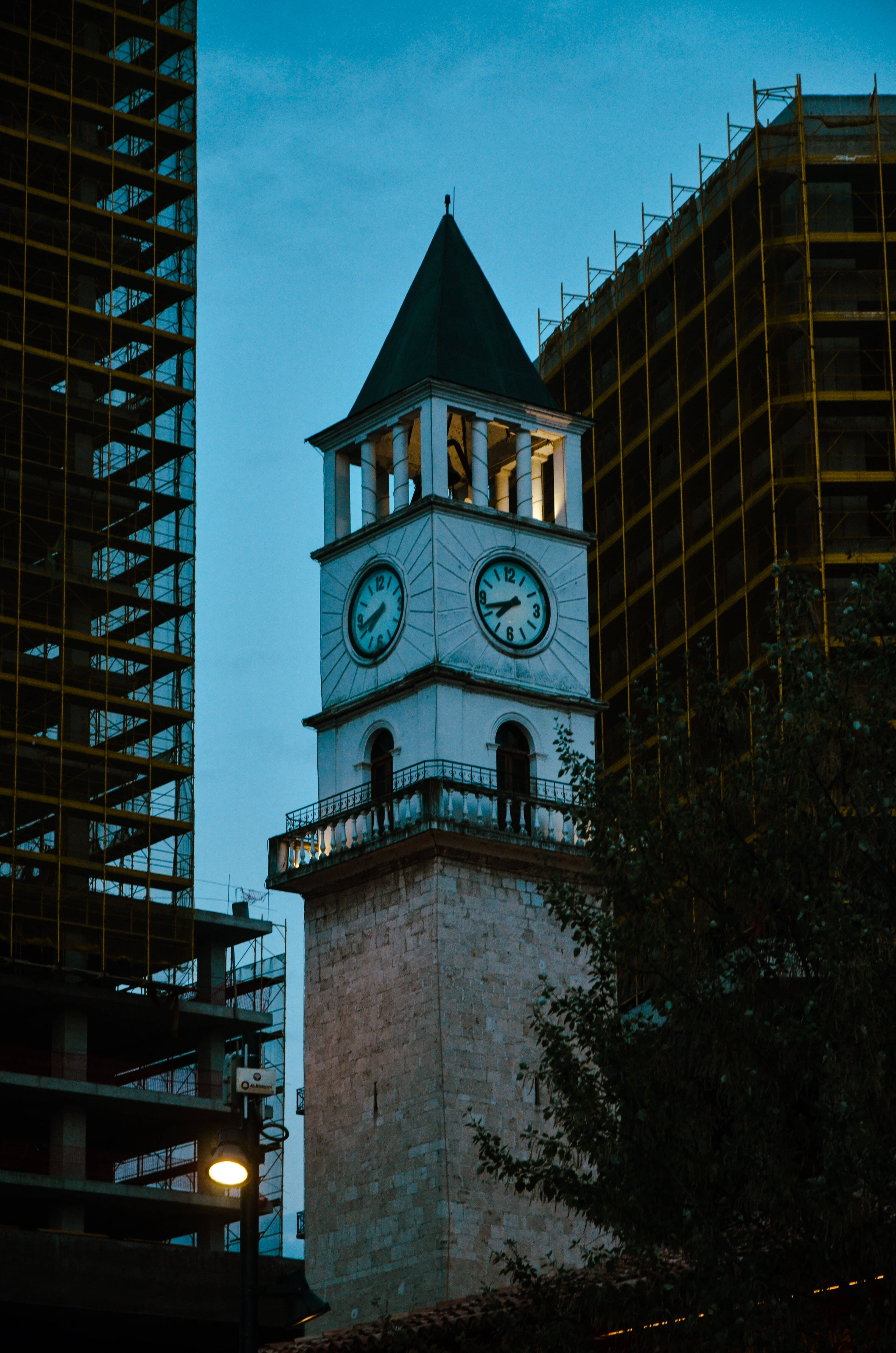Albania 桌布 壁紙 — 一座歷史悠久的石造鐘樓，帶有發光的鐘面，矗立在現代建築之間，背景是暮色天空。