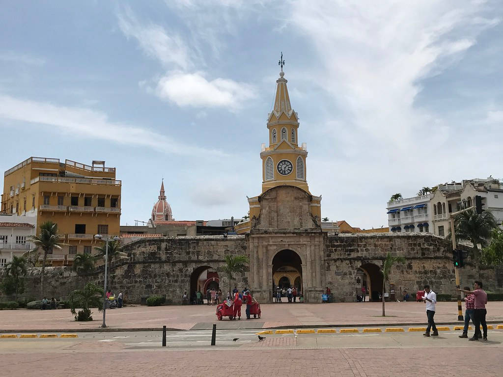 Colombia 桌布 壁紙 — 一座歷史悠久的石造鐘樓，帶有黃色尖頂，聳立在繁忙的城市廣場上，天空多雲。