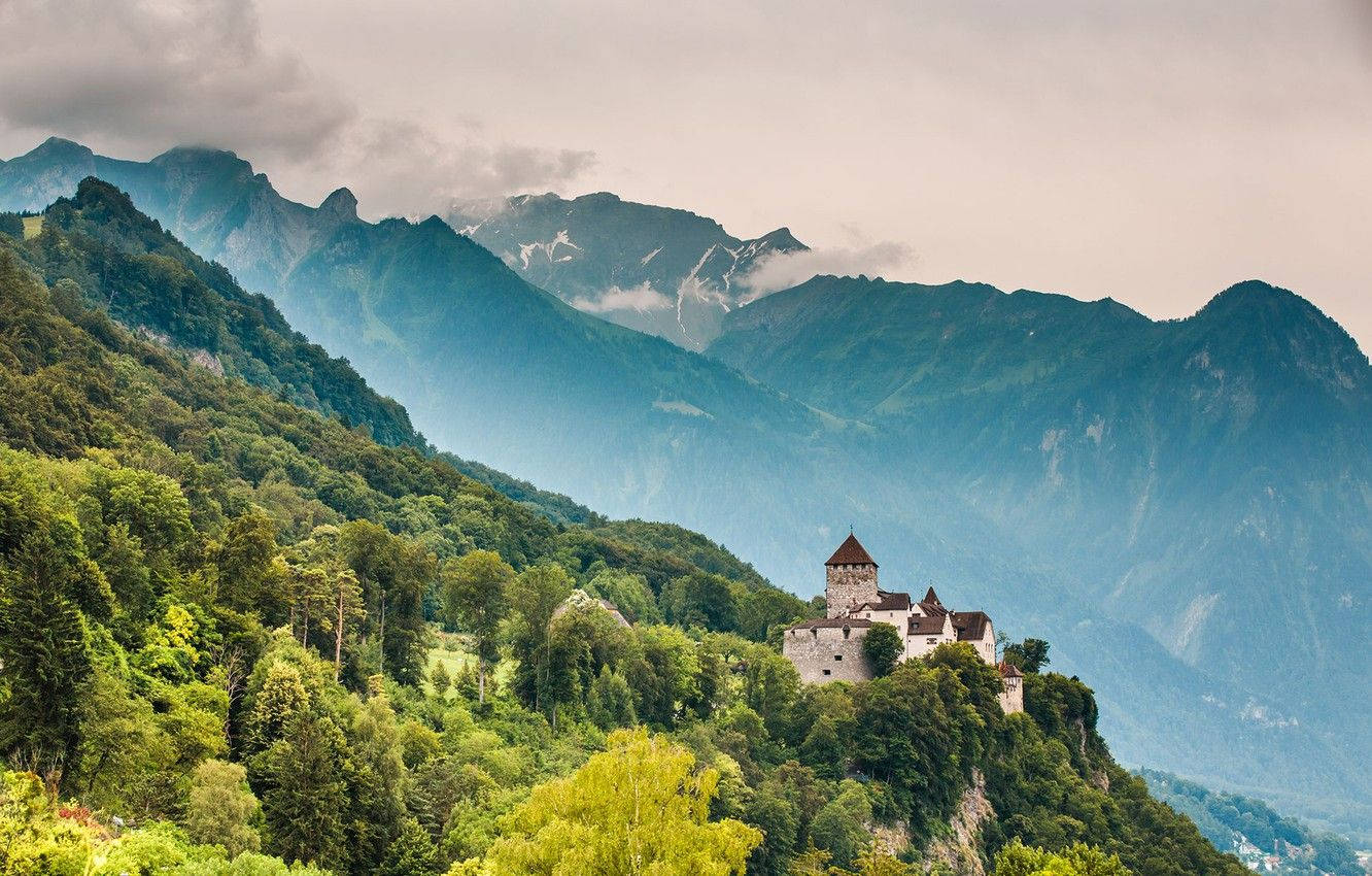 Liechtenstein 桌布 壁紙 — 一座歷史悠久的石頭城堡坐落在森林覆蓋的懸崖上，俯瞰著多雲天空下霧氣瀰漫的山脈。