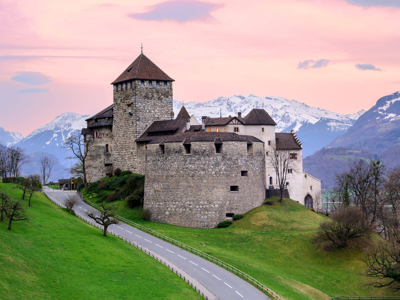 Liechtenstein 桌布 壁紙 — 一座歷史悠久的石頭城堡坐落在長滿青草的山丘上，一條蜿蜒的道路通向城堡，背景是白雪覆蓋的山脈，在柔和的粉紅色日落天空下。