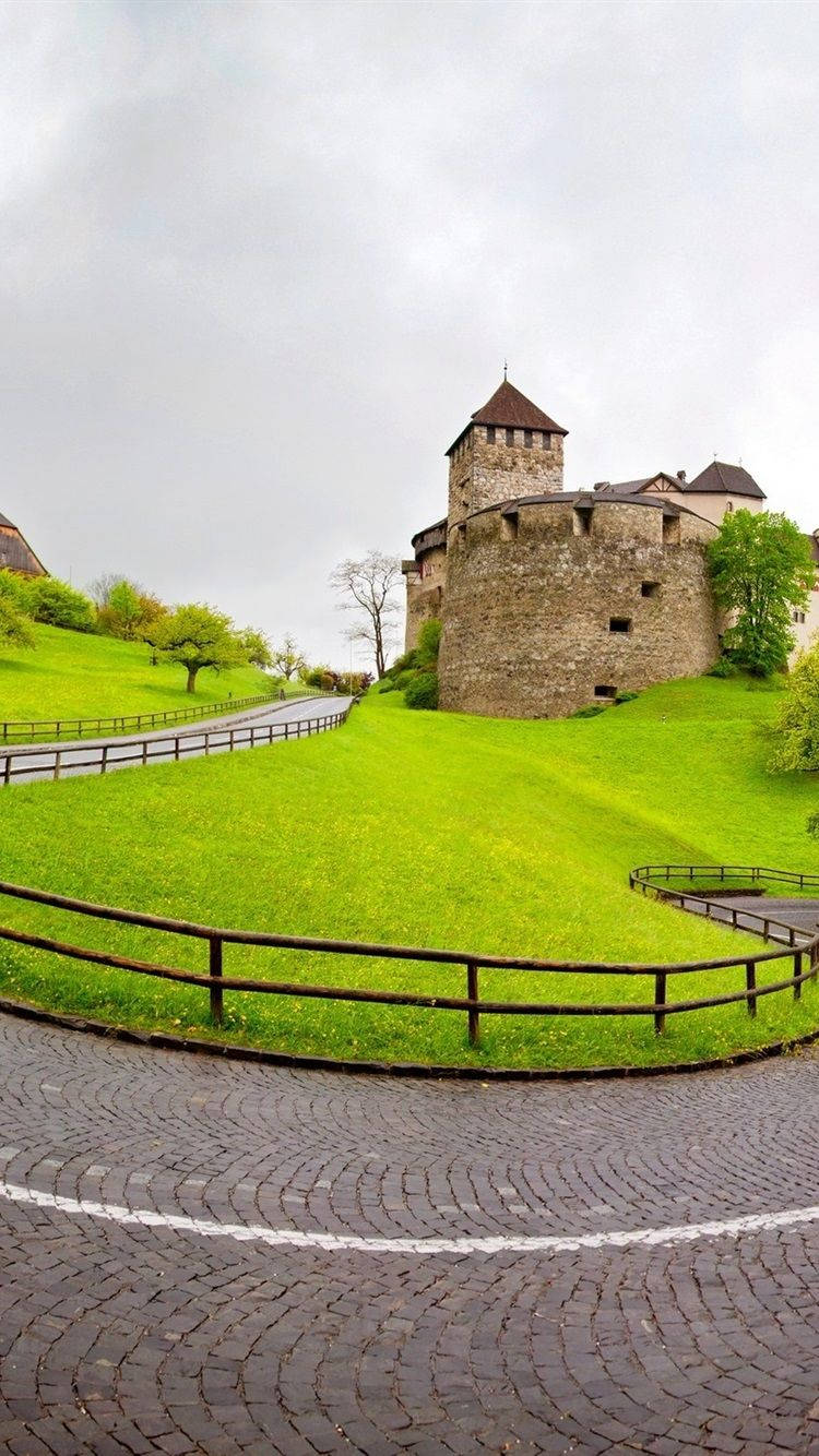 Liechtenstein 桌布 壁紙 — 一座歷史悠久的石頭城堡坐落在鬱鬱蔥蔥的綠色山頂上，從蜿蜒的鵝卵石路上觀看，天空多雲。