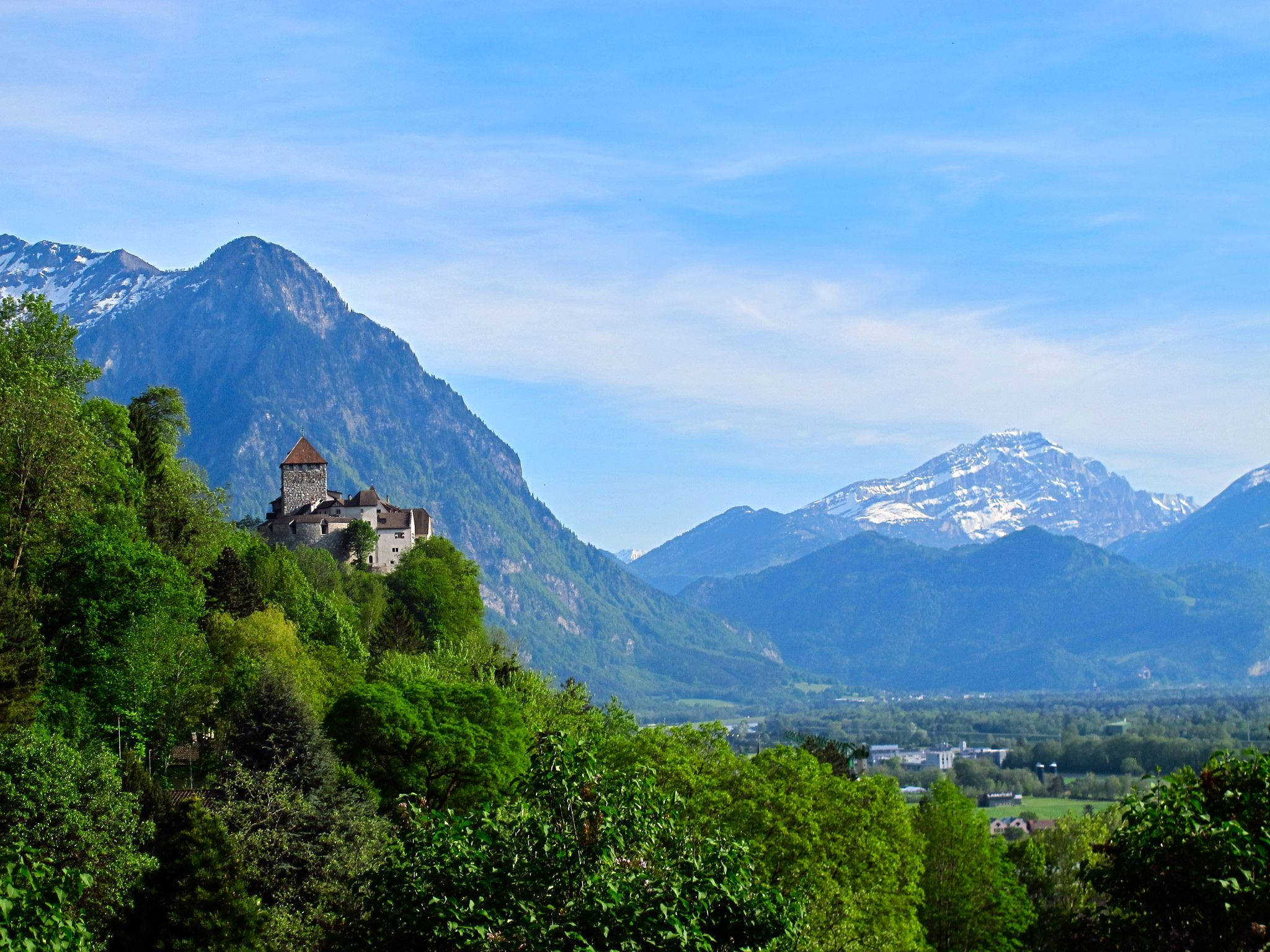 Liechtenstein 桌布 壁紙 — 一座歷史悠久的石頭城堡坐落在鬱鬱蔥蔥的綠色山坡上，背景是白雪覆蓋的山峰在清澈的藍天下雄偉地聳立。