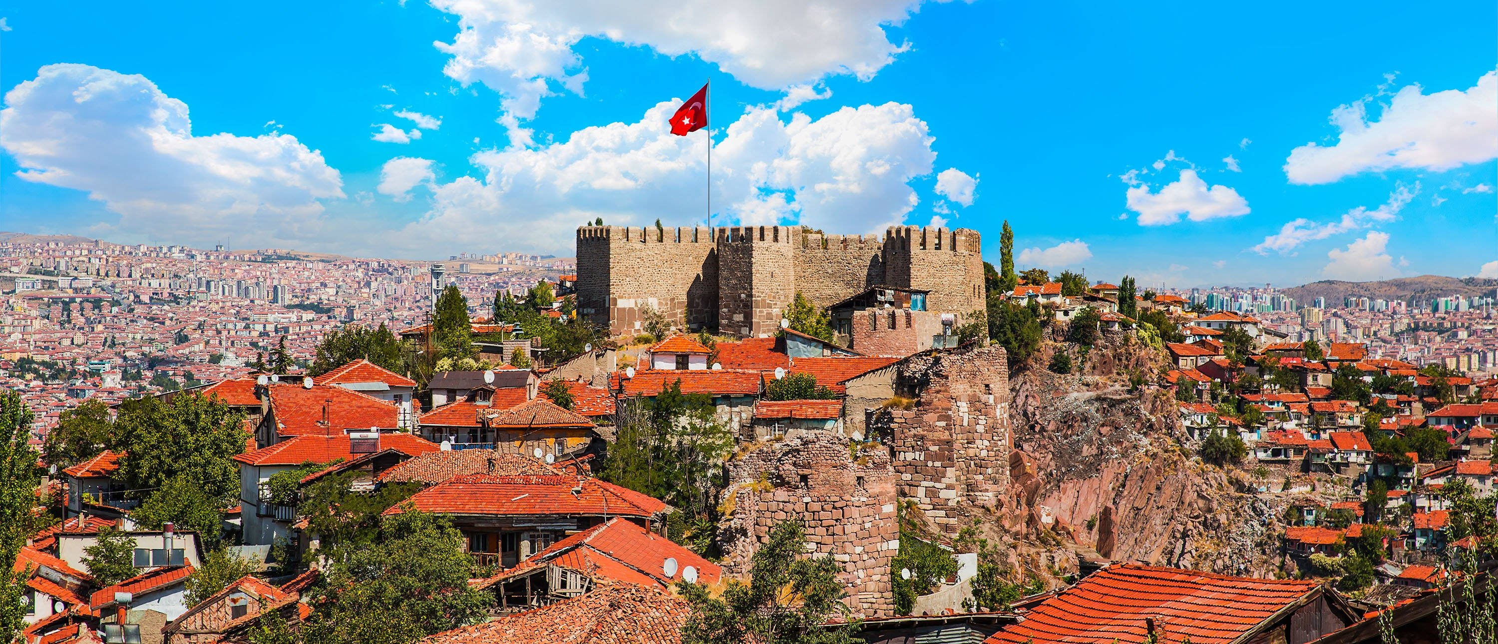 Ankara 桌布 壁紙 — 一座歷史悠久的石頭堡壘，頂上飄揚著紅旗，矗立在岩石山丘上，俯瞰著一座擁有陶土屋頂的廣闊城市，天空湛藍。