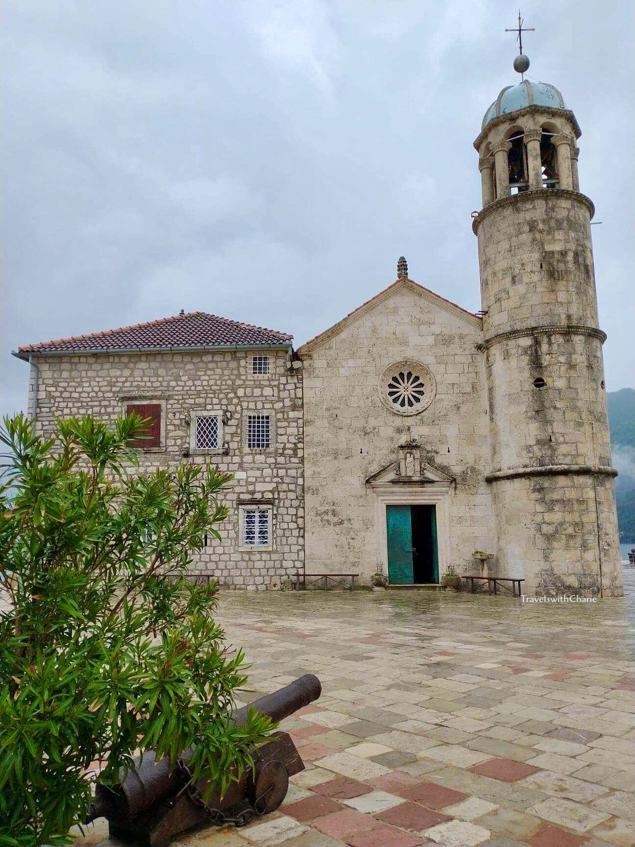 Montenegro 桌布 壁紙 — 一座歷史悠久的石頭教堂，帶有鐘樓和綠色門，矗立在陰天之下，旁邊有一門大砲和茂密的綠植。