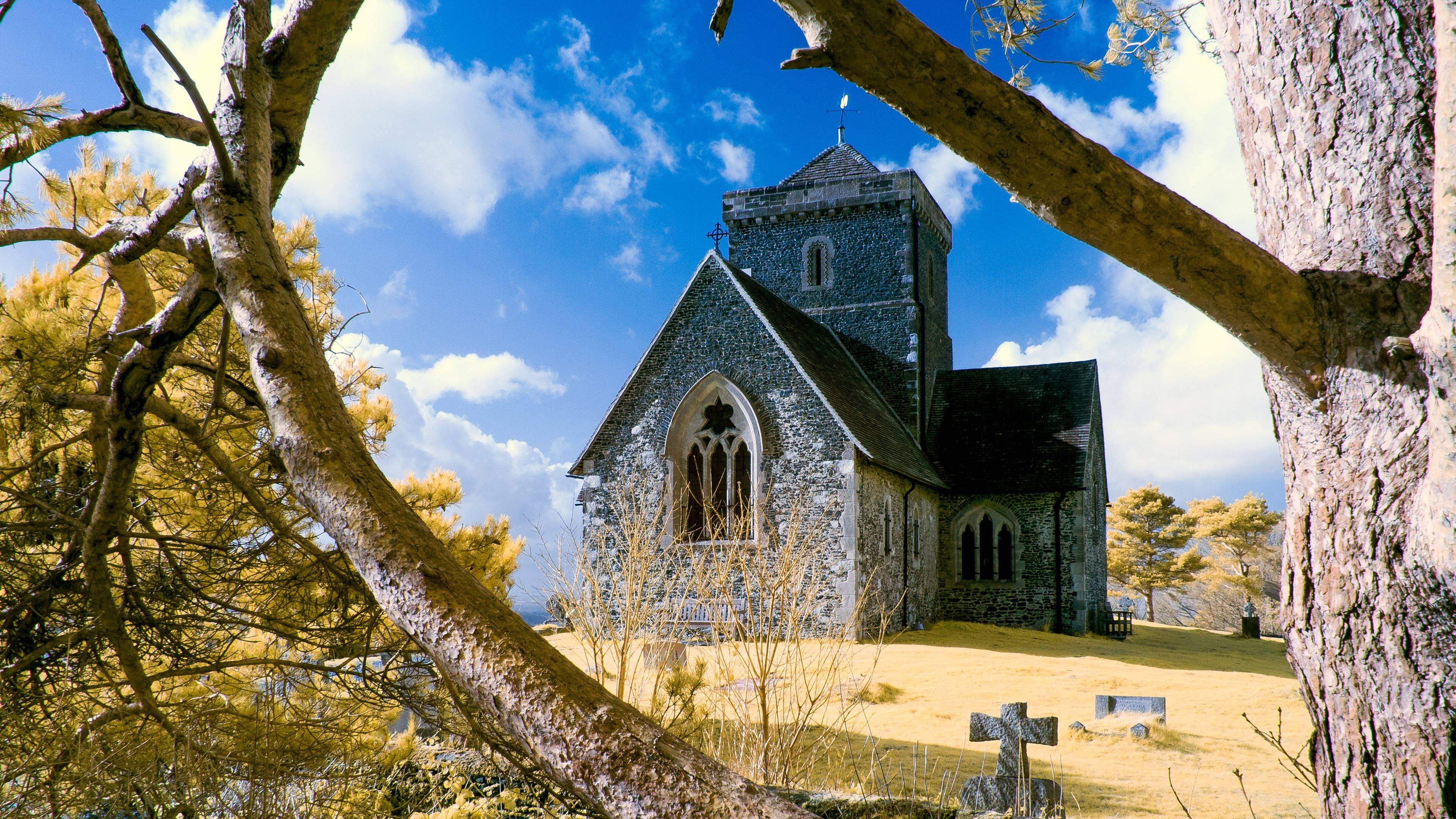 England 桌布 壁紙 — 一座歷史悠久的石頭教堂，被樹枝框住，背景是明亮的藍天和蓬鬆的雲朵。