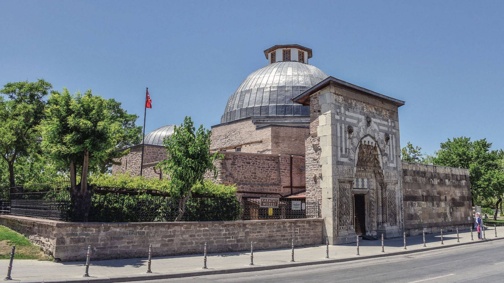 Konya 桌布 壁紙 — 一座歷史悠久的石頭清真寺，擁有大型銀色圓頂和拱形入口，矗立在晴朗的藍天下，周圍環繞著綠樹。
