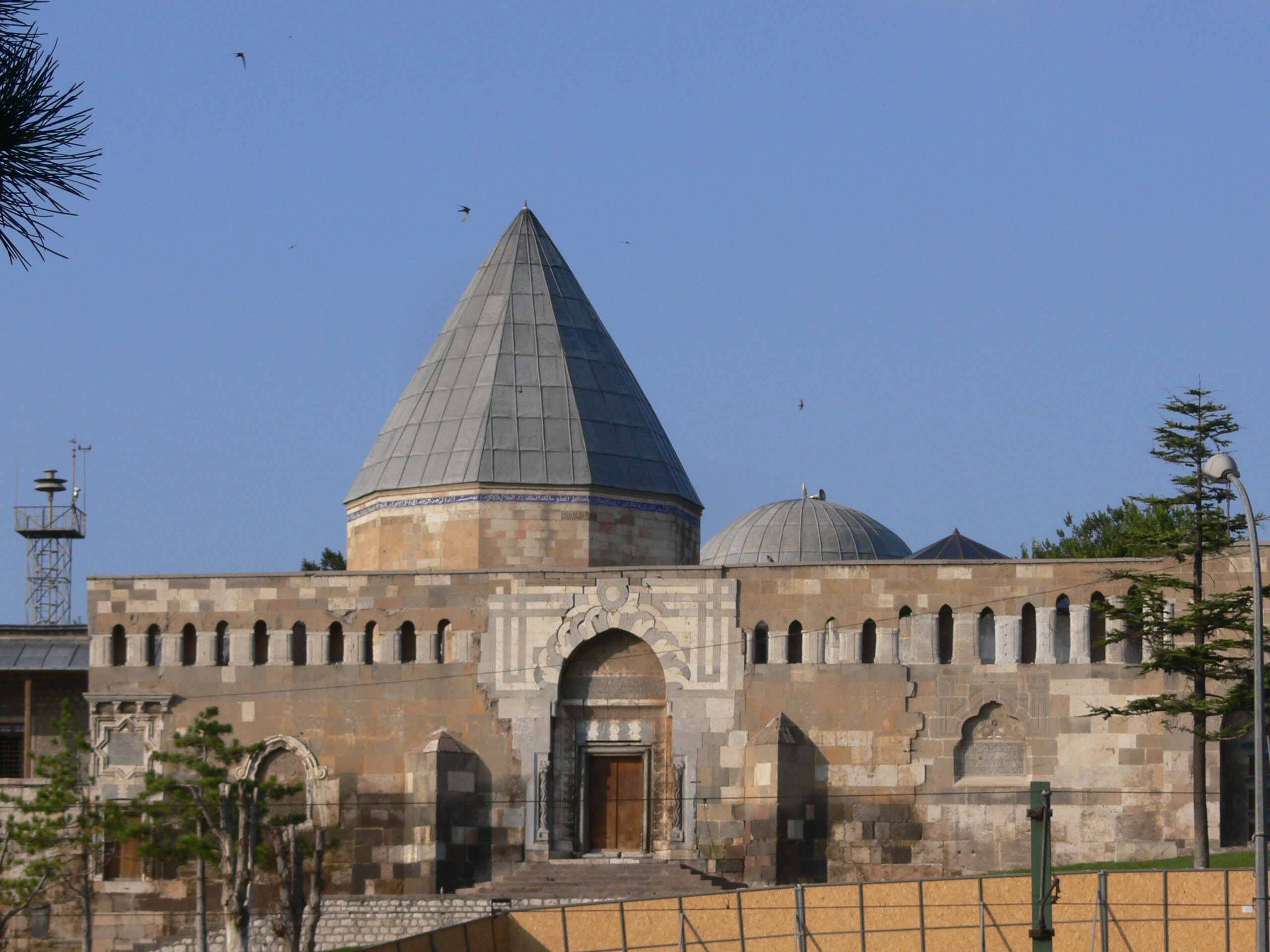 Konya 桌布 壁紙 — 一座歷史悠久的石頭清真寺，擁有高高的圓錐形屋頂和拱形入口，矗立在晴朗的藍天下，頭頂有零星鳥兒飛過。