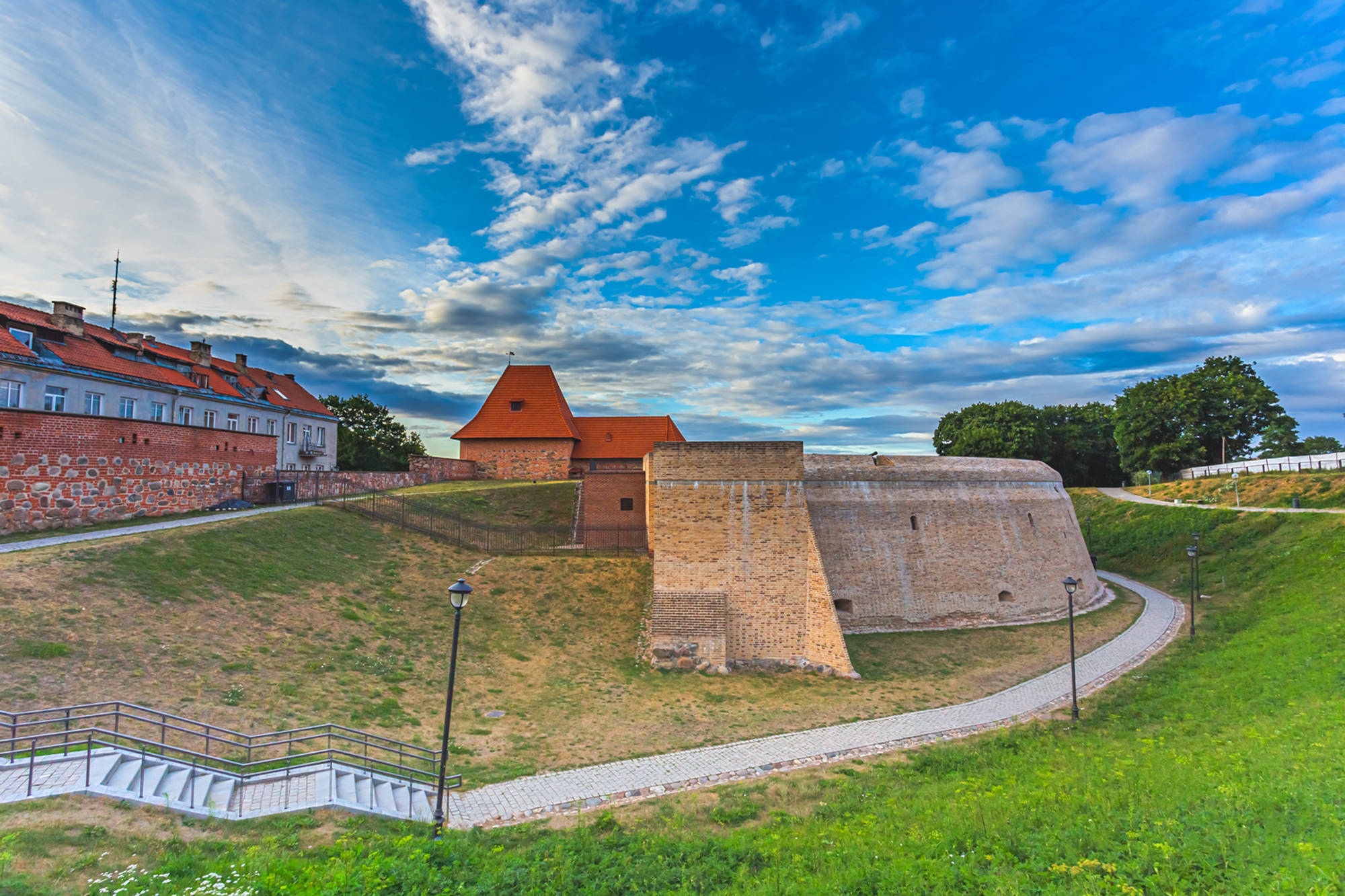 Vilnius 桌布 壁紙 — 一座歷史悠久的磚砌堡壘，紅色屋頂矗立在長滿青草的山丘上，天空湛藍，雲朵輕盈。
