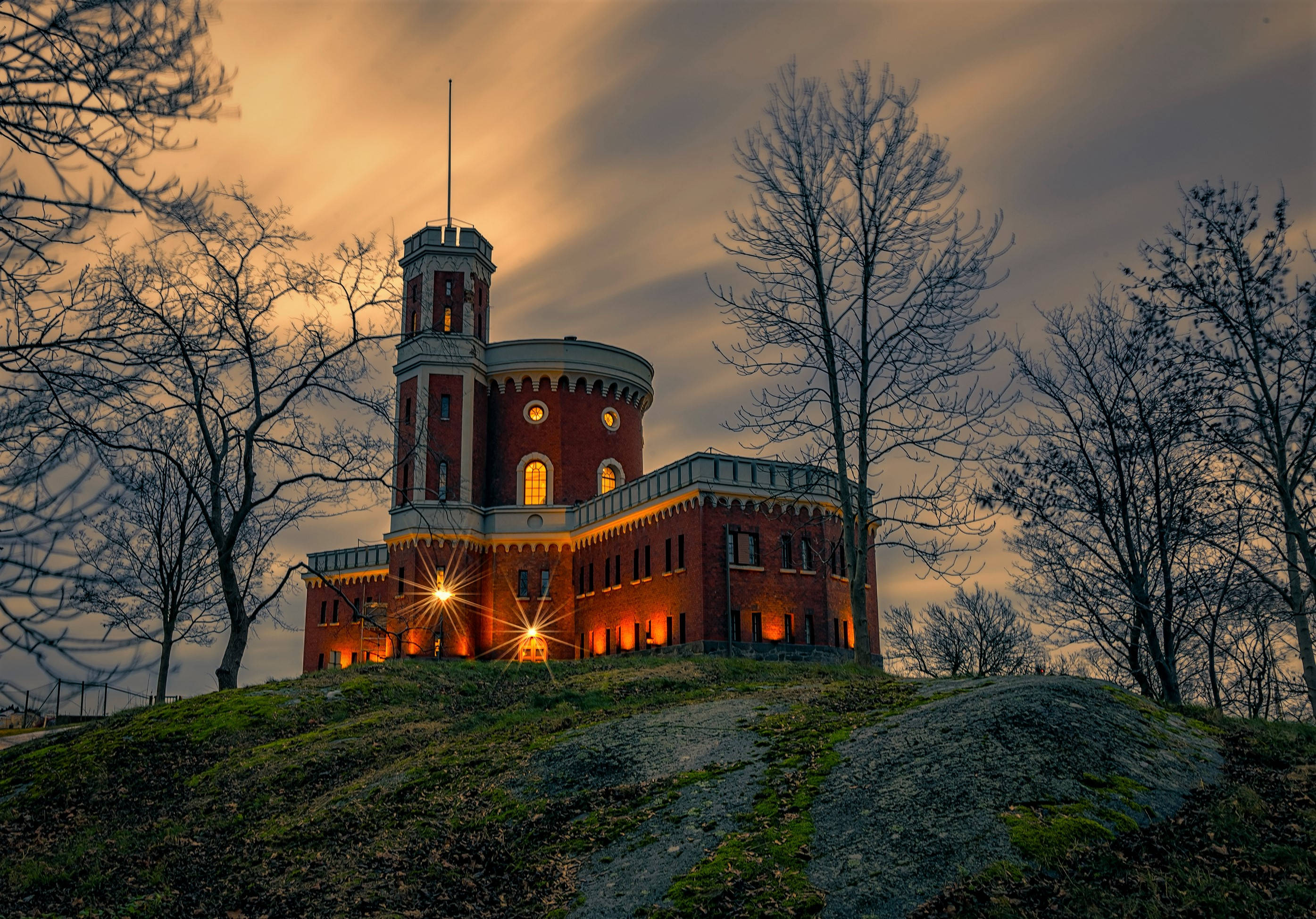 Sweden 桌布 壁紙 — 一座歷史悠久的紅磚城堡坐落在長滿青草的山丘上，在光禿禿的樹木襯托下，於戲劇性的暮色天空中溫暖地發光。