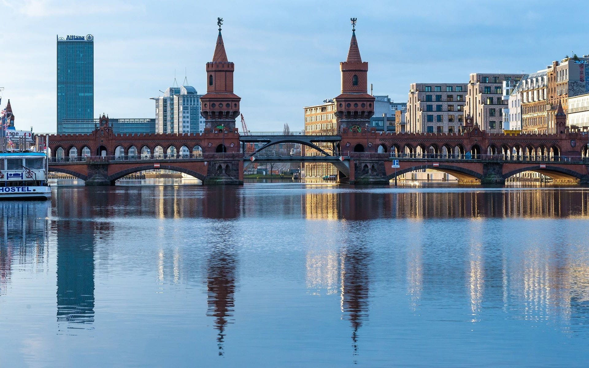Berlin 桌布 壁紙 — 一座歷史悠久的紅磚橋，帶有雙塔，橫跨平靜的河流，倒映著現代摩天大樓和清澈的藍天。