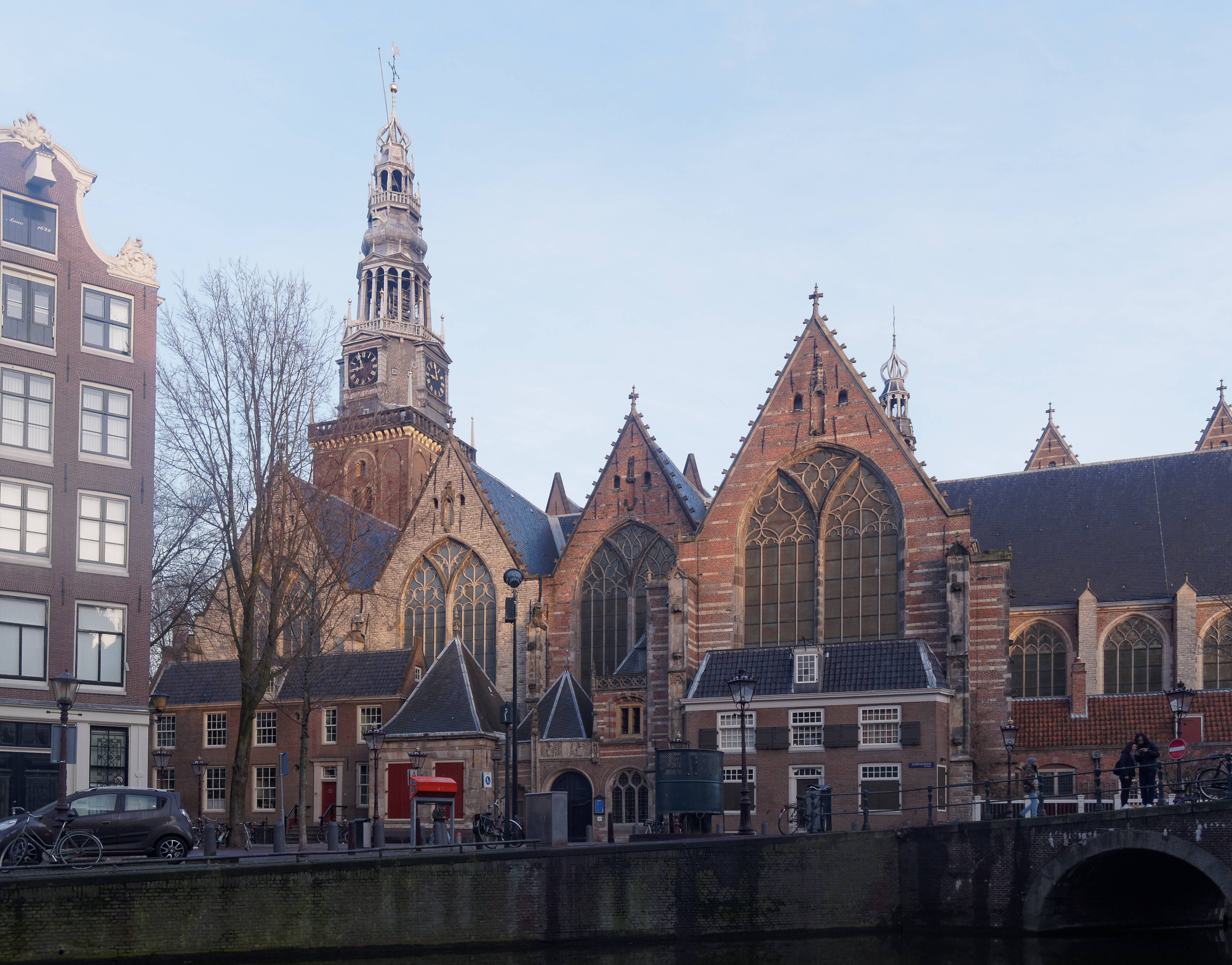 Amsterdam 桌布 壁紙 — 一座歷史悠久的荷蘭教堂，擁有高高的鐘樓，矗立在運河旁，天空清澈湛藍，附近有光禿禿的樹木。