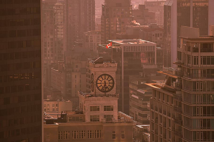 Clock 桌布 壁紙 — 一座歷史悠久的鐘樓矗立在沐浴著溫暖金色光芒的摩天大樓之間，上方飄揚著加拿大國旗。