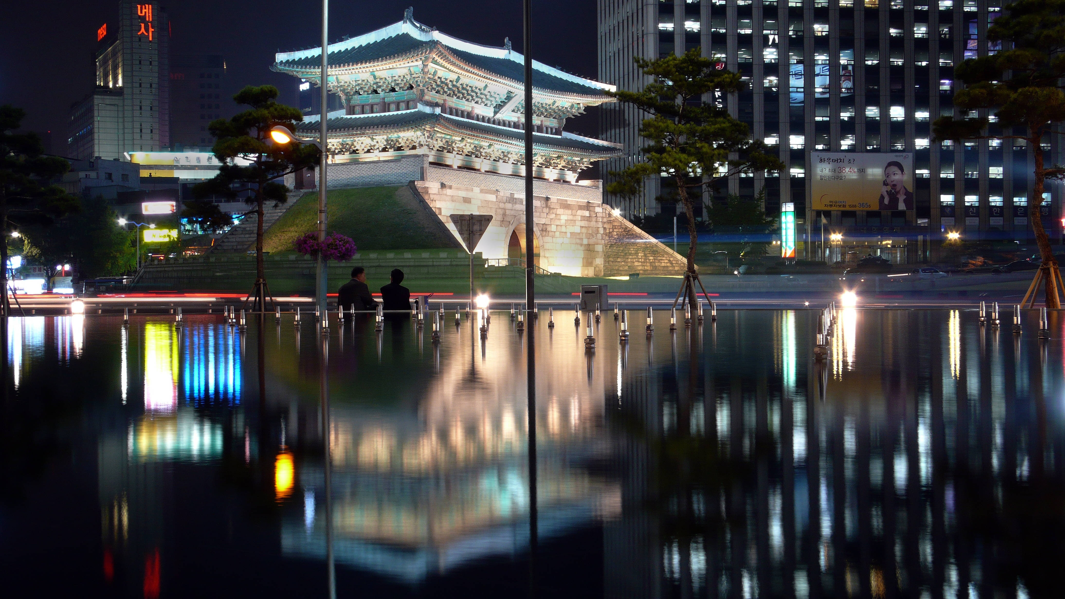 南韓 桌布 壁紙 — 一座歷史悠久的韓國城門在夜晚發光，燈光倒映在平靜的水池中，背景是模糊的城市燈光。