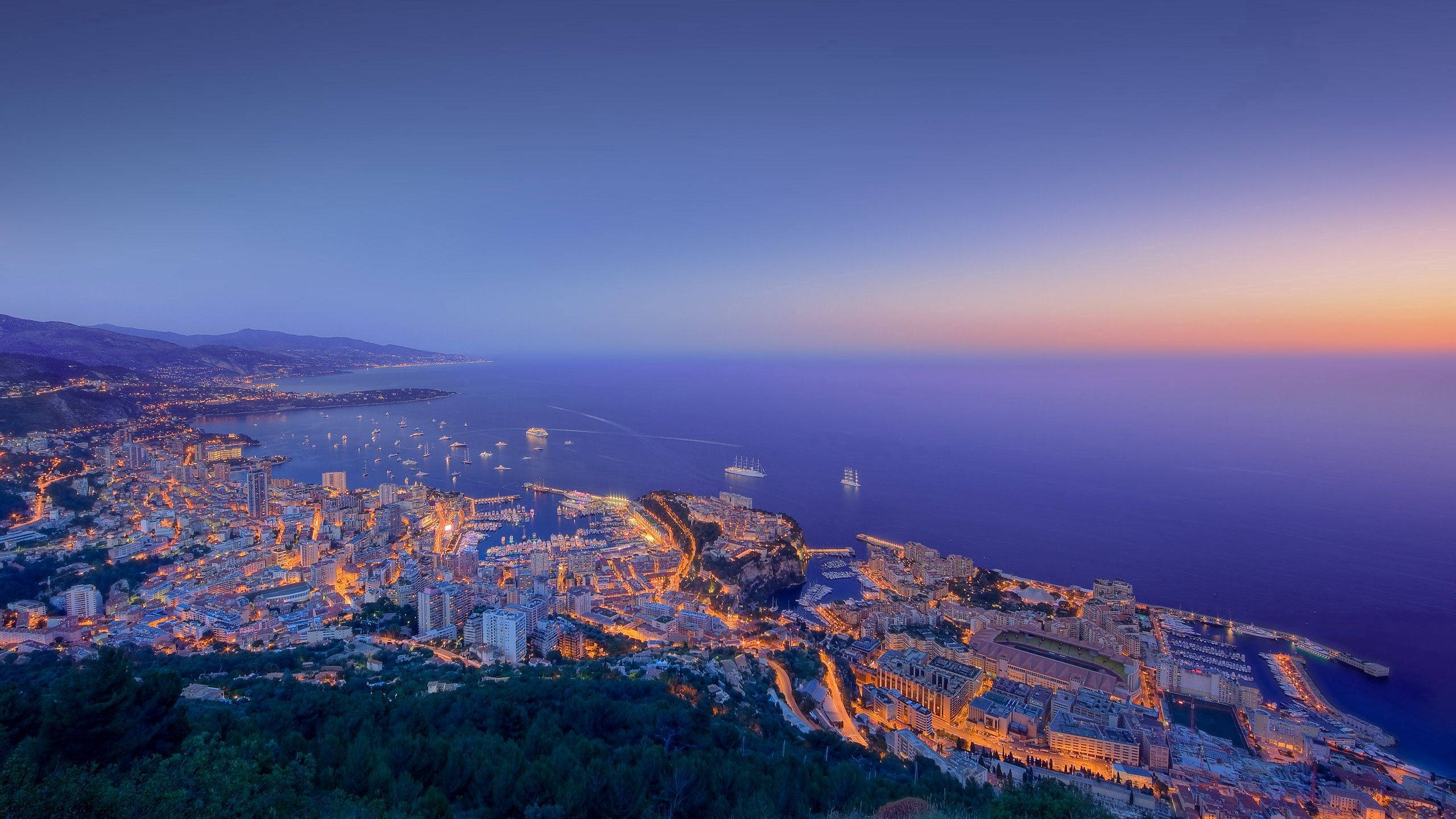 Monaco 桌布 壁紙 — 一座沿海城市在暮色天空下閃耀著金色燈光，船隻點綴在平靜的海面上。