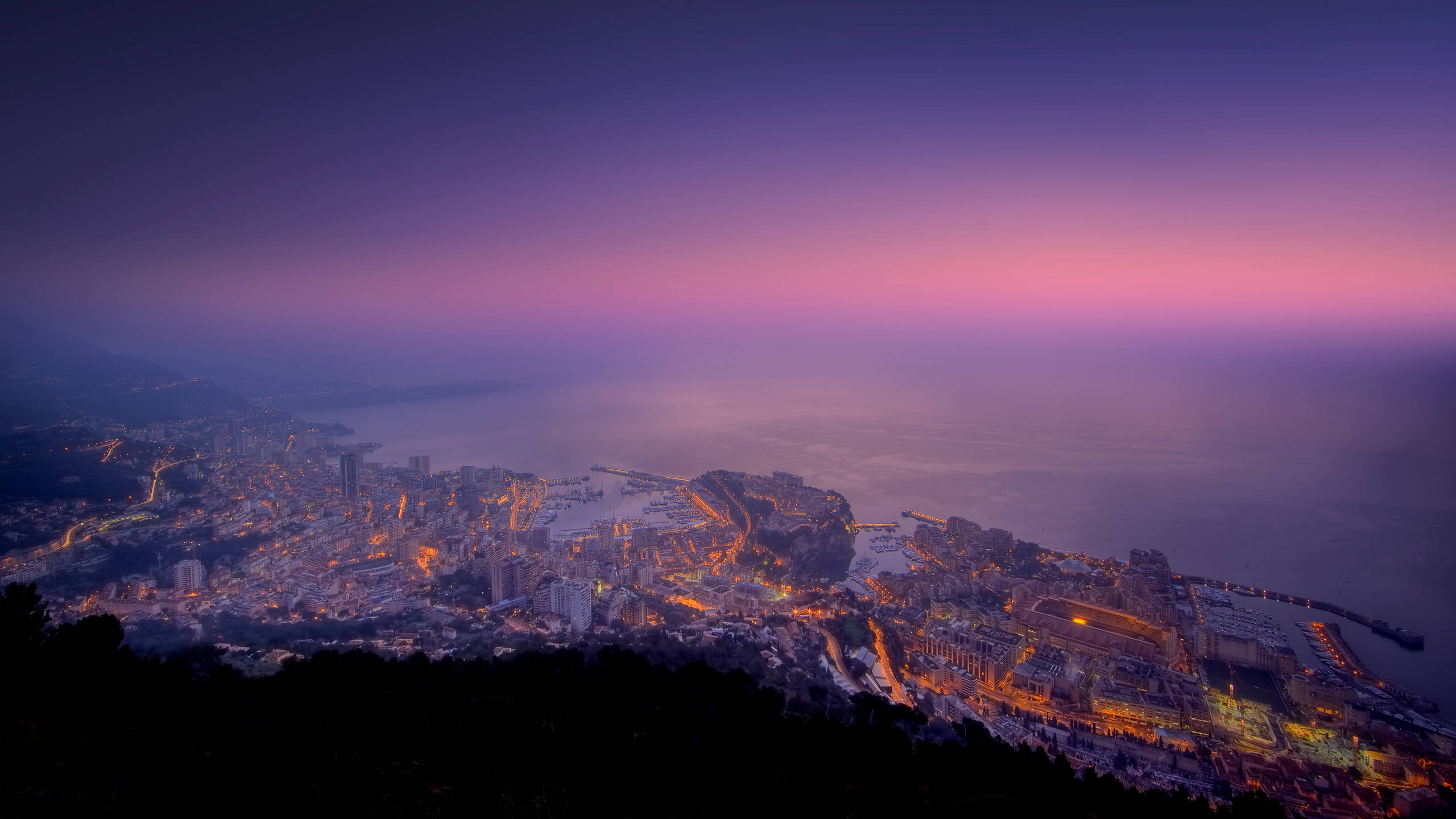 Monaco 桌布 壁紙 — 一座沿海城市在深紫色和粉紅色的暮色天空下，散發著溫暖的光芒。