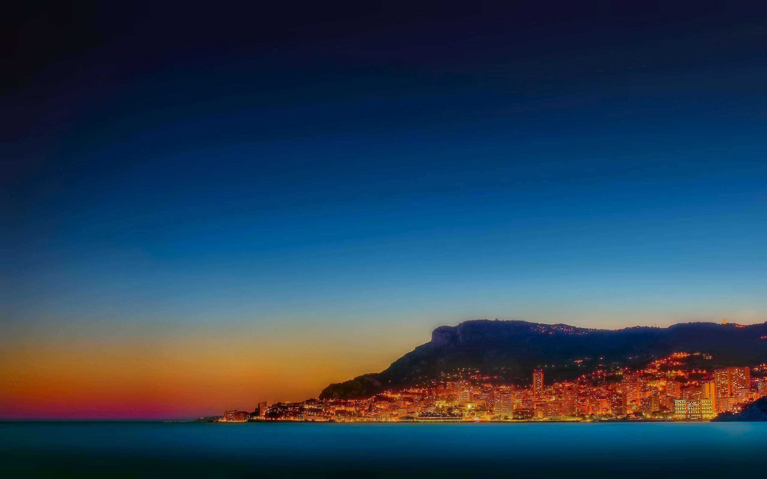 Monaco 桌布 壁紙 — 一座沿海城市在溫暖的燈光下閃耀，暮色天空從深藍色過渡到地平線附近的火橙色。