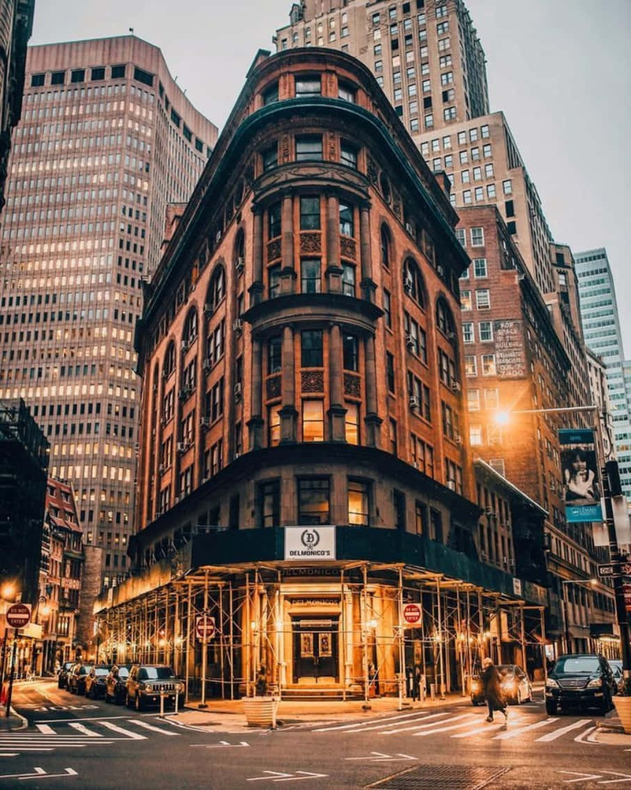 Nyc Aesthetic 桌布 壁紙 — 一座溫暖照明的歷史棕色石頭角落建築，矗立在繁華城市街道上高聳的現代摩天大樓之間，正值黃昏時分。