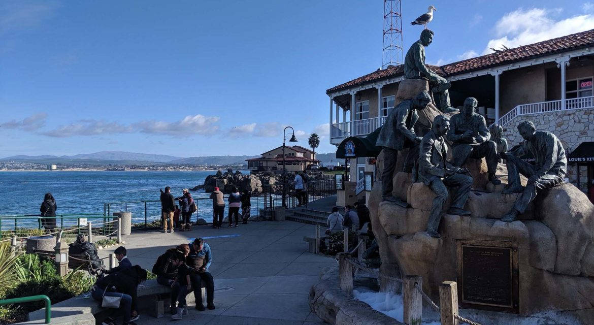 Statue Aesthetic 桌布 壁紙 — 一座漁夫銅像棲息在海邊步道旁的石頭上，人們在晴朗的藍天下放鬆。