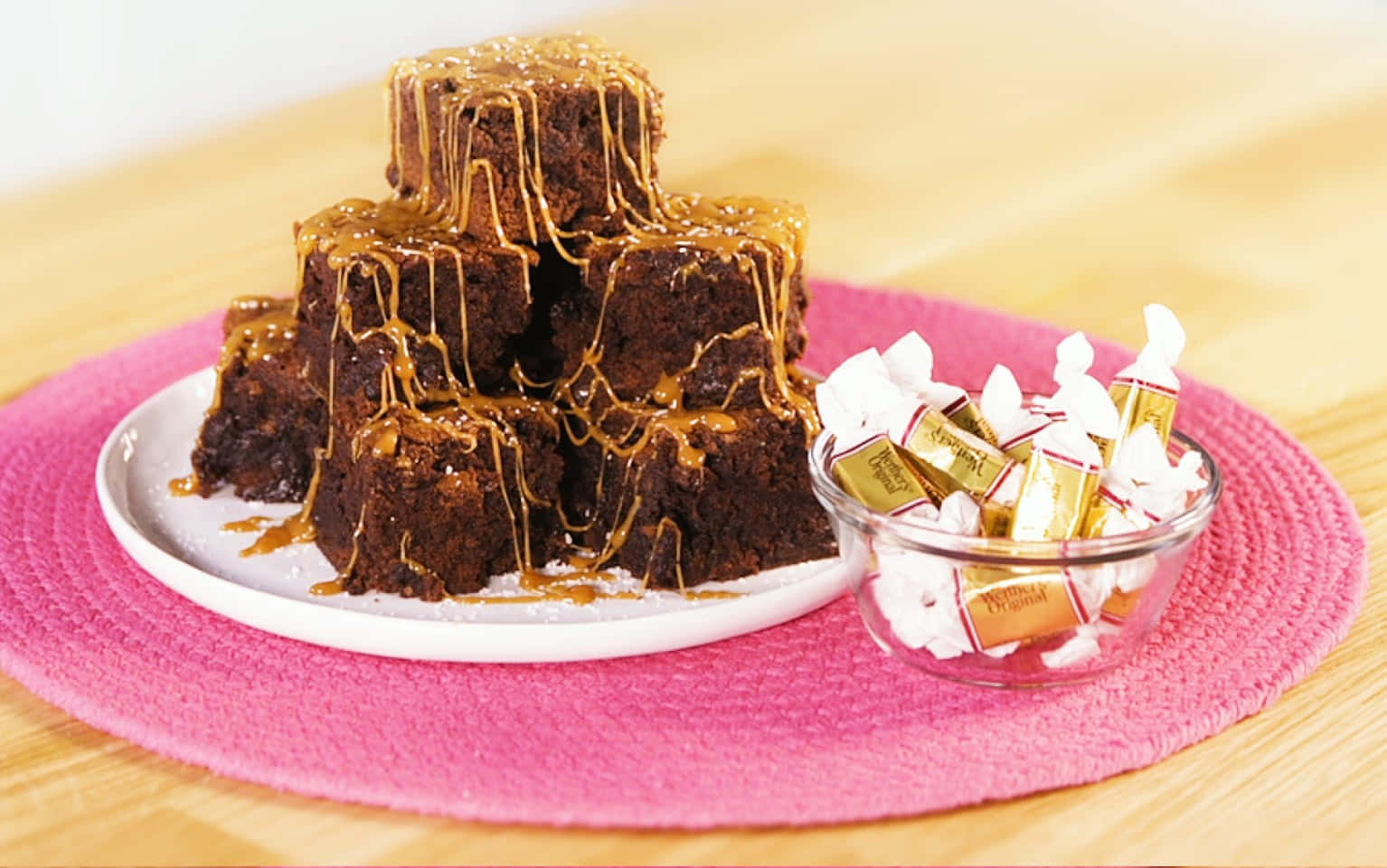 Caramel Brownie 桌布 壁紙 — 一座濃郁巧克力布朗尼塔，淋上焦糖醬，旁邊放著一碗包裝巧克力，放在粉紅色餐墊上。