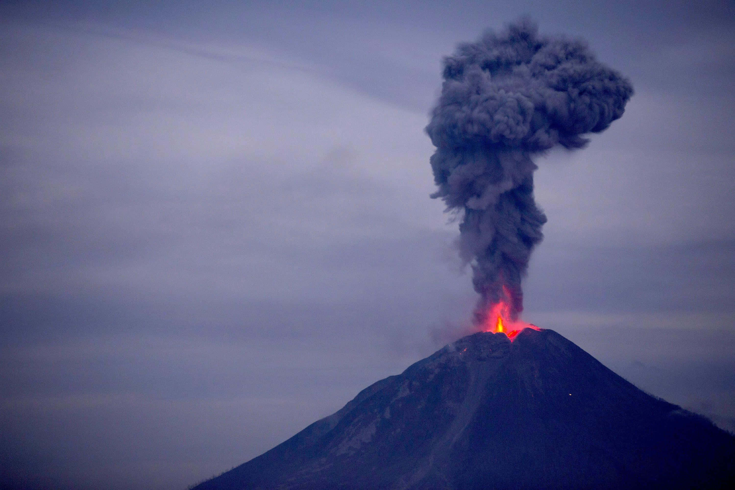 Volcano 桌布 壁紙 — 一座火山爆發，噴出熾熱的熔岩和巨大的黑煙柱，映襯著暮色天空。