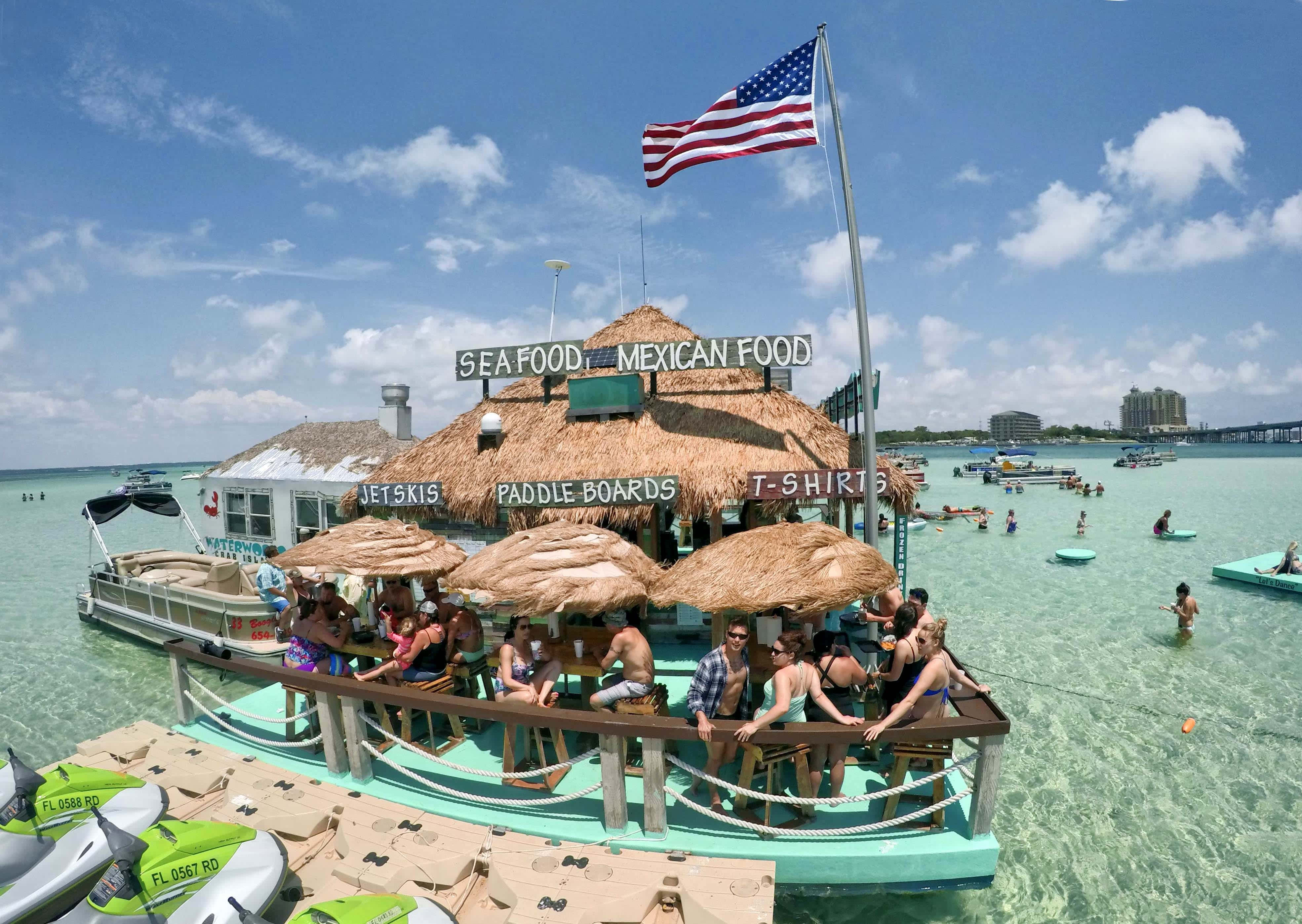 Crab Island 桌布 壁紙 — 一座熱鬧的漂浮海鮮和墨西哥美食攤位，位於碧綠色的水上，顧客在茅草傘下，附近有水上摩托車。