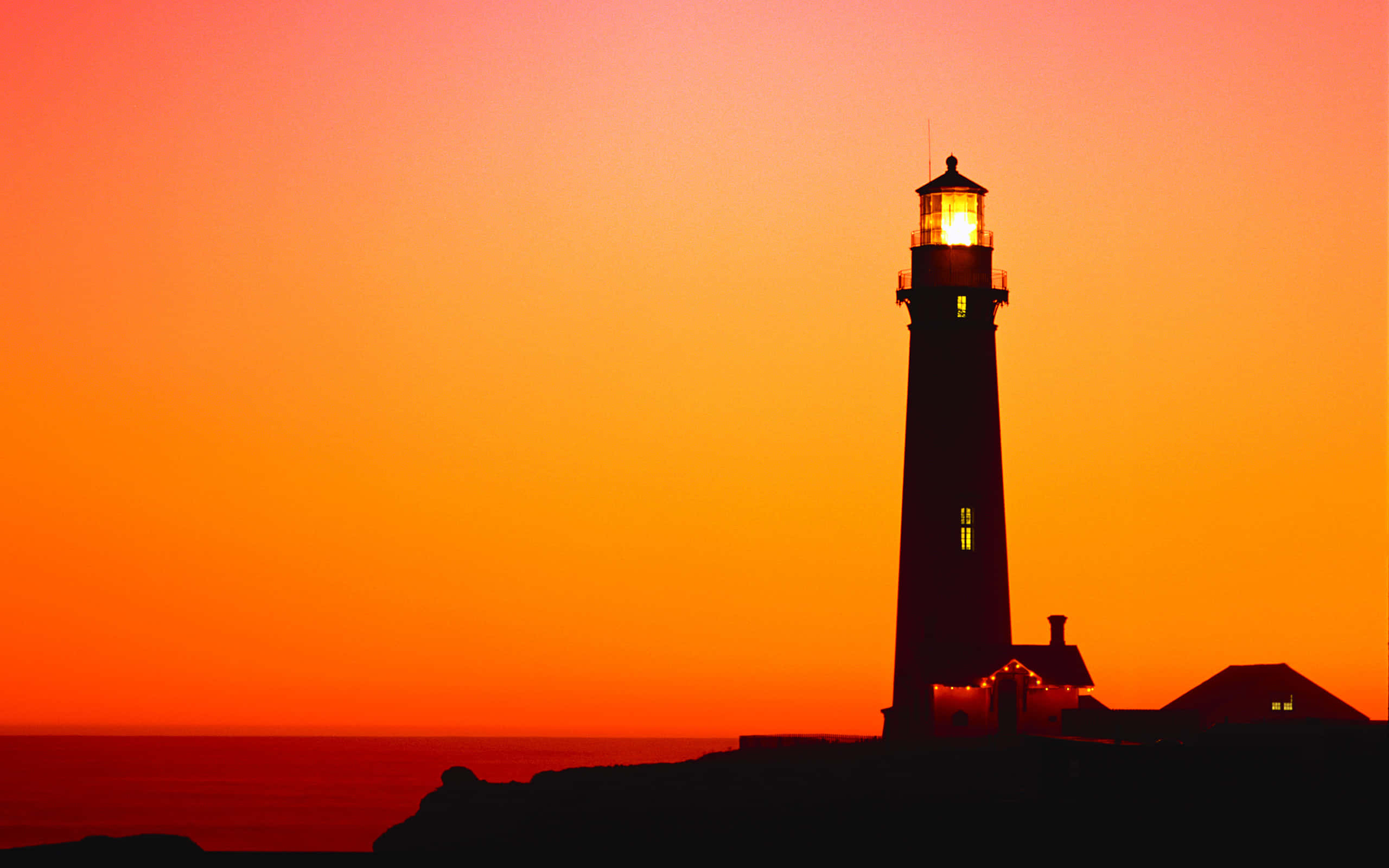 Lighthouse 桌布 壁紙 — 一座燈塔在海洋上熾熱的橙色日落中呈現剪影，其燈塔溫暖地發光。