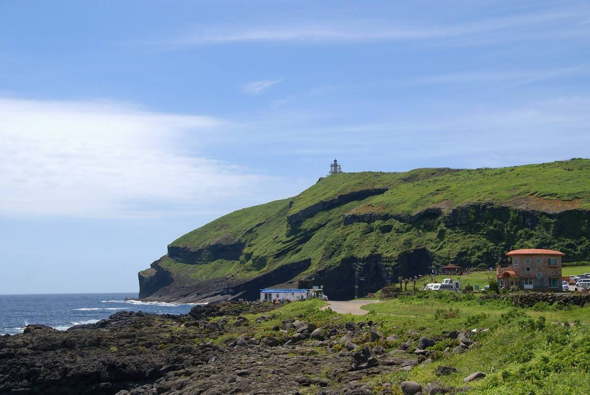 Jeju 桌布 壁紙 — 一座燈塔坐落在鬱鬱蔥蔥的綠色懸崖上，俯瞰海洋，下方有蜿蜒的道路和色彩繽紛的建築，在明亮的藍天下。