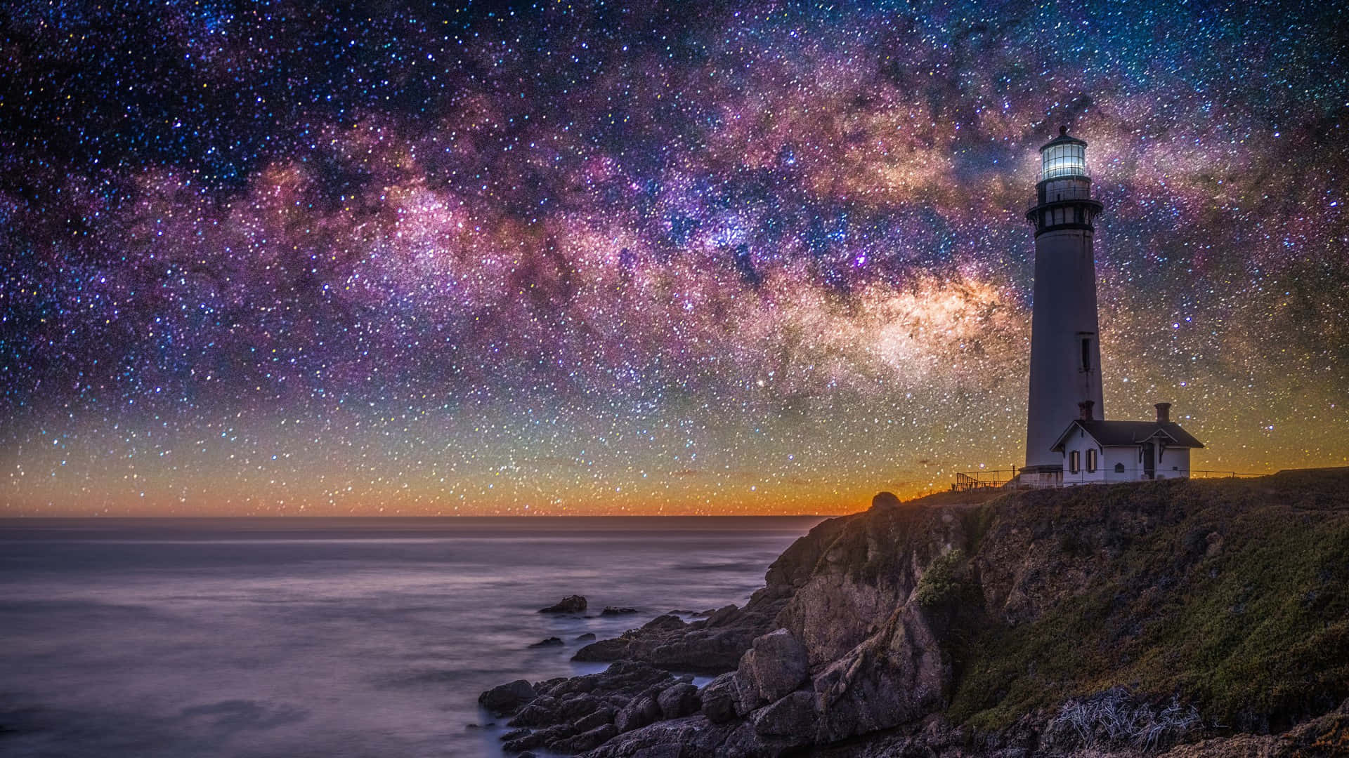 Colorful Sky 桌布 壁紙 — 一座燈塔矗立在岩石懸崖上，在暮色中，一條充滿活力的紫色和粉紅色銀河拱跨平靜的海洋。
