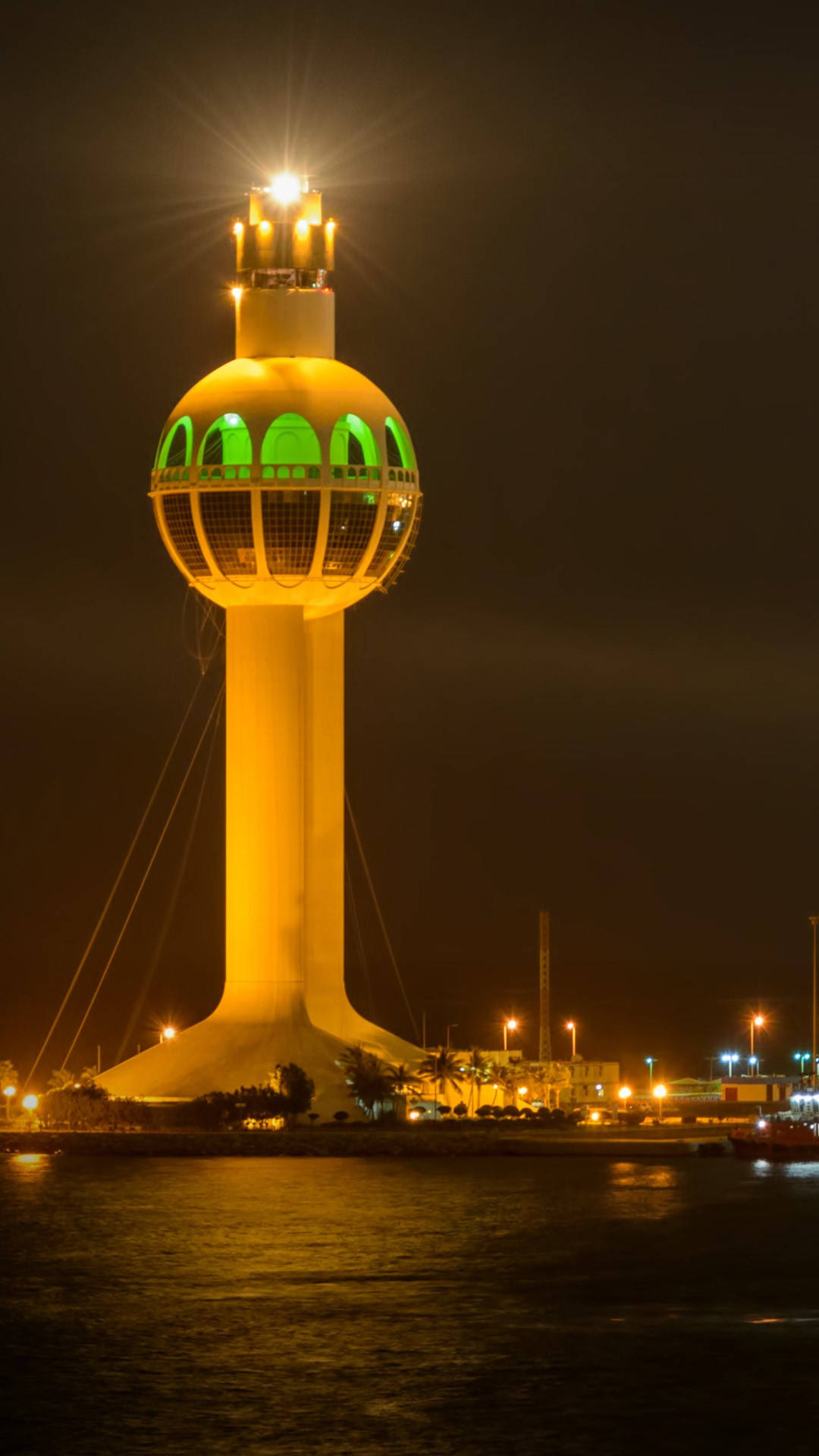 Saudi 桌布 壁紙 — 一座燈火通明的黃色塔樓，頂部有發光的綠色圓頂，夜晚矗立在水面上，在黑暗的水面投下倒影。