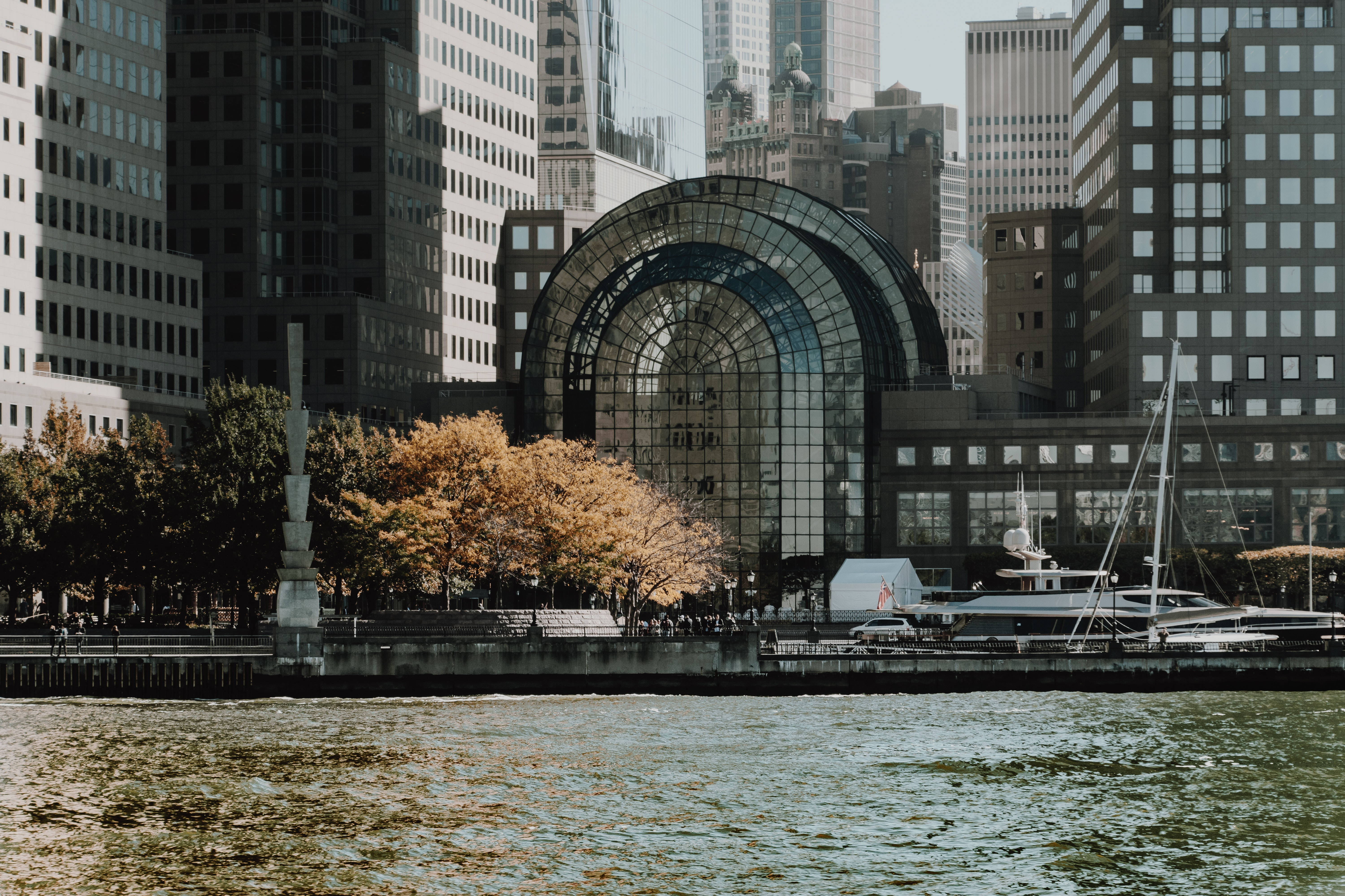 Nyc Aesthetic 桌布 壁紙 — 一座玻璃圓頂建築矗立在城市高樓大廈之間，旁邊有一條河流，河邊有秋天的樹木，附近還停泊著一艘帆船。