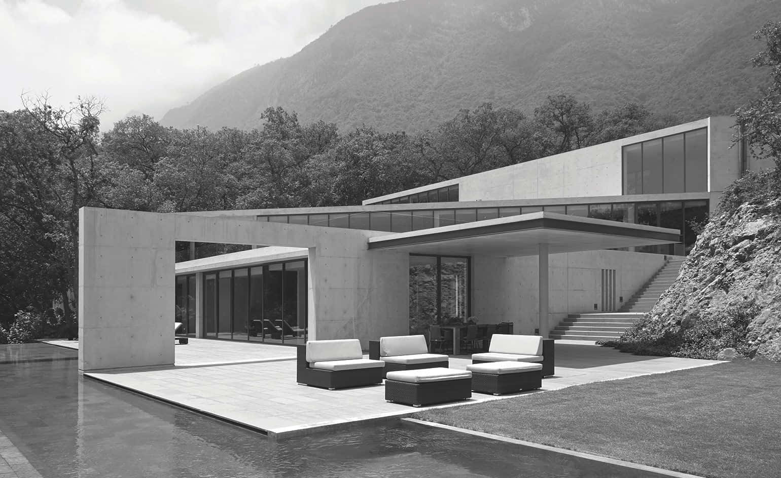 Black And White Architecture 桌布 壁紙 — 一座現代主義混凝土房屋，配有玻璃牆和戶外座位，位於游泳池旁，背景是單色調的森林覆蓋的山脈。