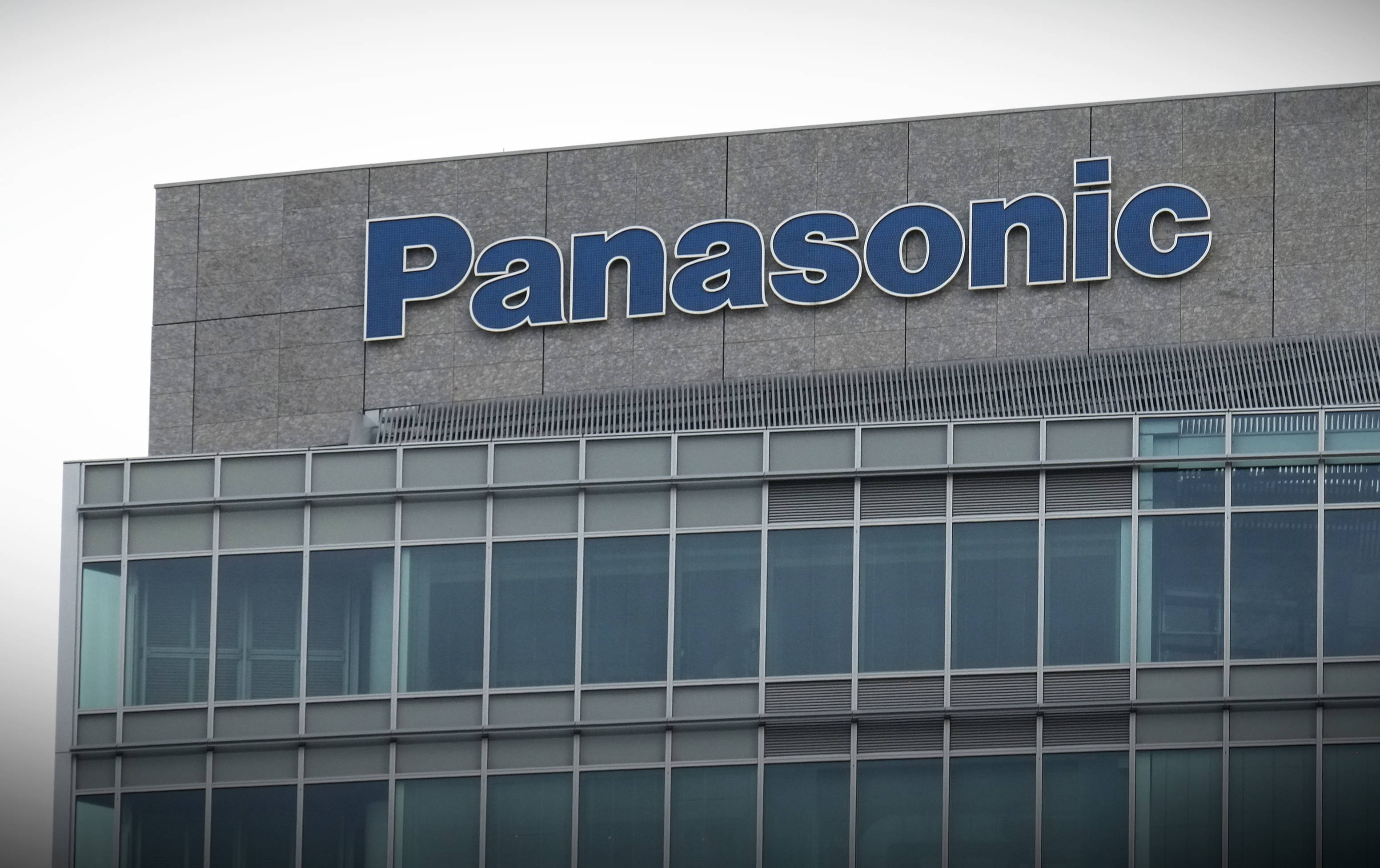 Panasonic 桌布 壁紙 — 一座現代企業建築，灰色石牆上有大型藍色 Panasonic 標誌，天空多雲。