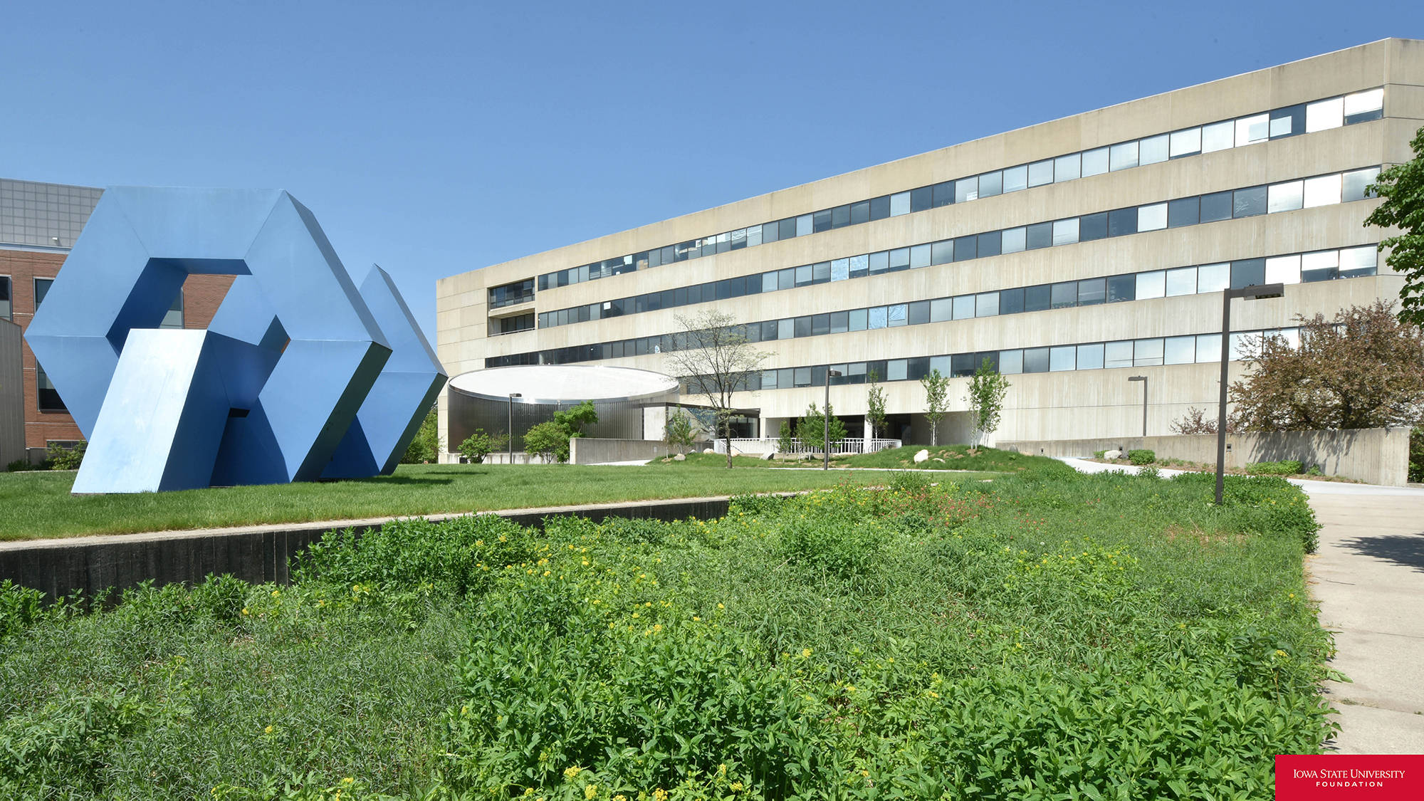 Building 桌布 壁紙 — 一座現代化大學建築，在晴朗藍天下，綠色景觀庭院中有一個大型藍色幾何雕塑。
