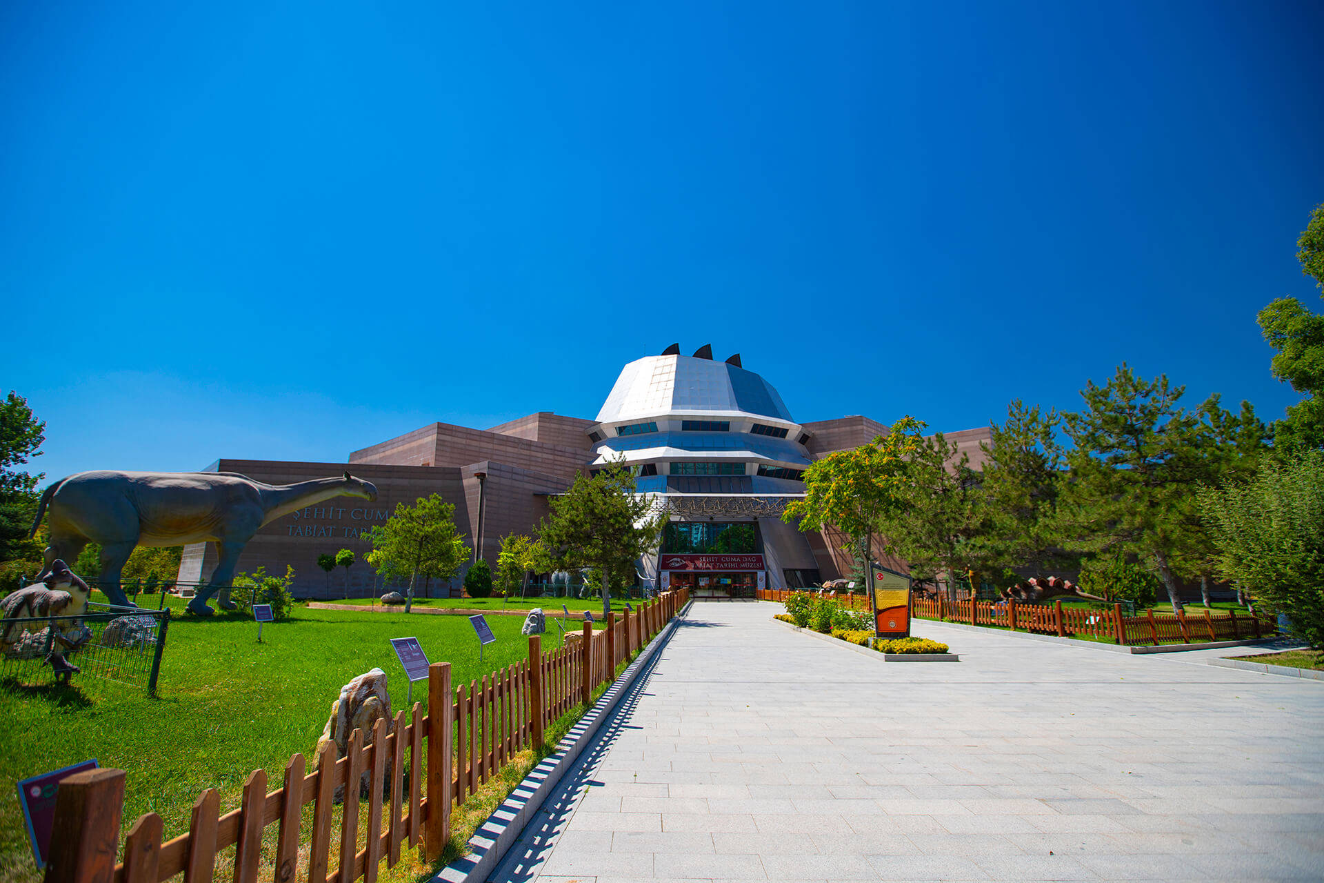 Ankara 桌布 壁紙 — 一座現代博物館，擁有白色圓頂和棕色牆壁，矗立在清澈的藍天下，兩旁有恐龍雕像和綠樹。