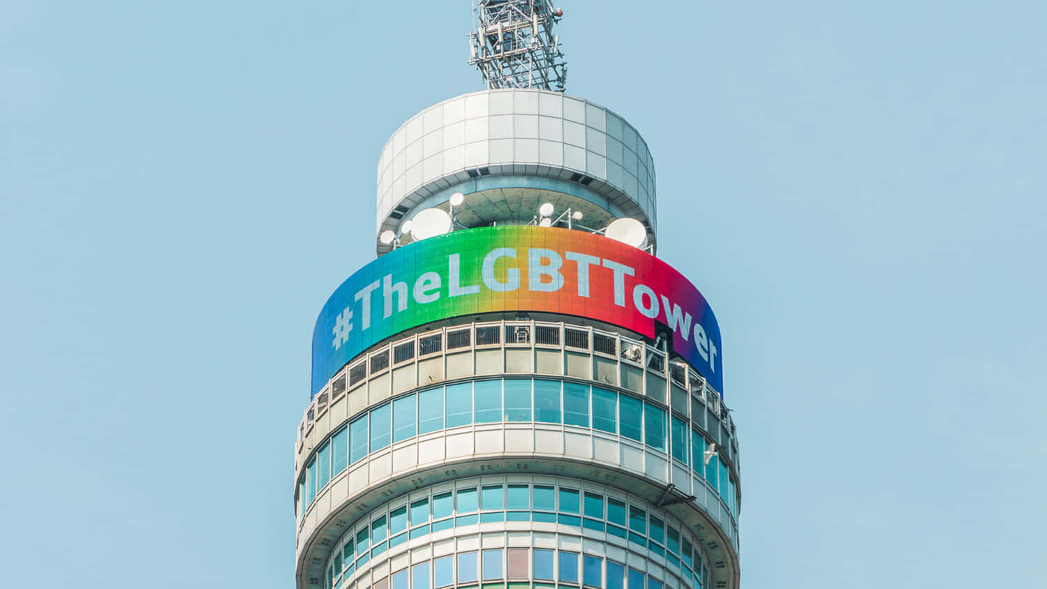 Blue Sky 桌布 壁紙 — 一座現代塔樓在清澈的藍天下展示帶有標籤 TheLGBTTower 的鮮豔彩虹橫幅。