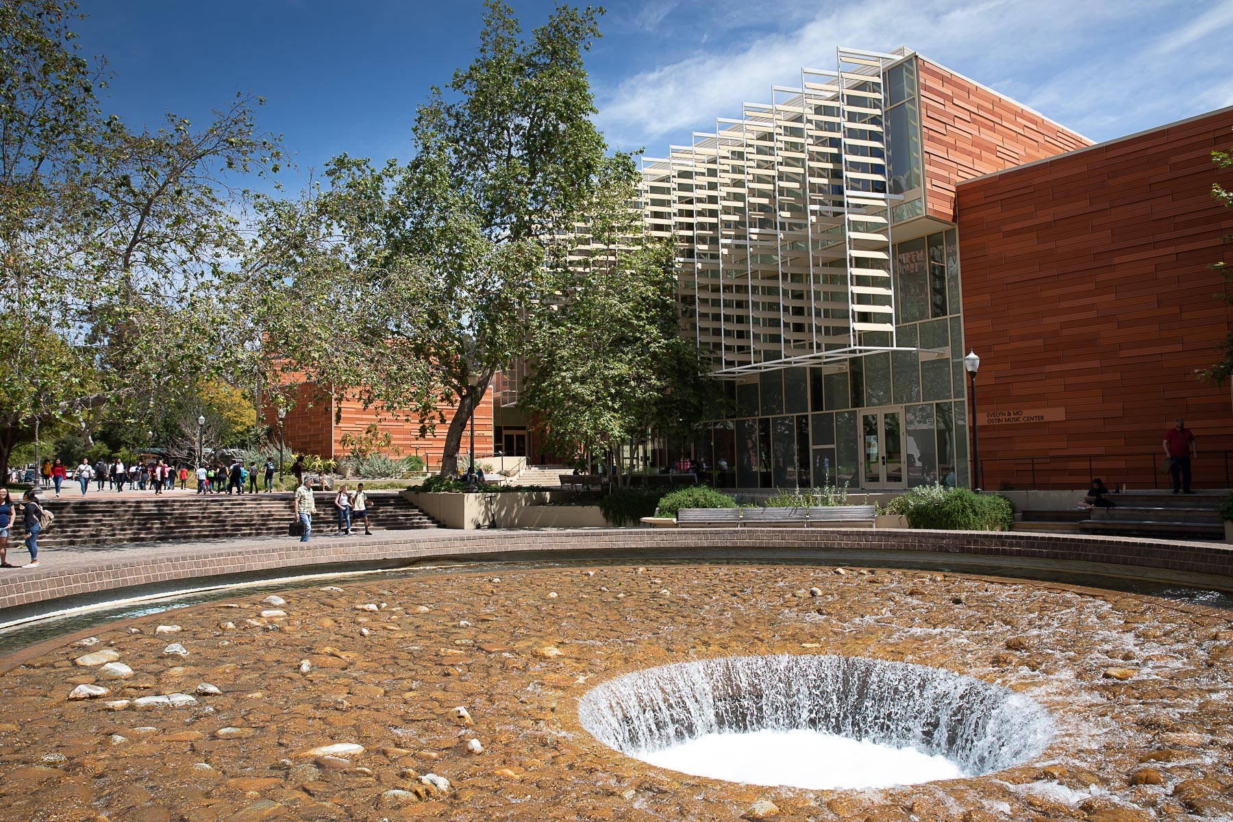 ucla 桌布 壁紙 — 一座現代大學建築，採用玻璃和赤陶外牆，矗立在圓形石噴泉後面，噴泉水流傾瀉而下，背景是清澈的藍天。