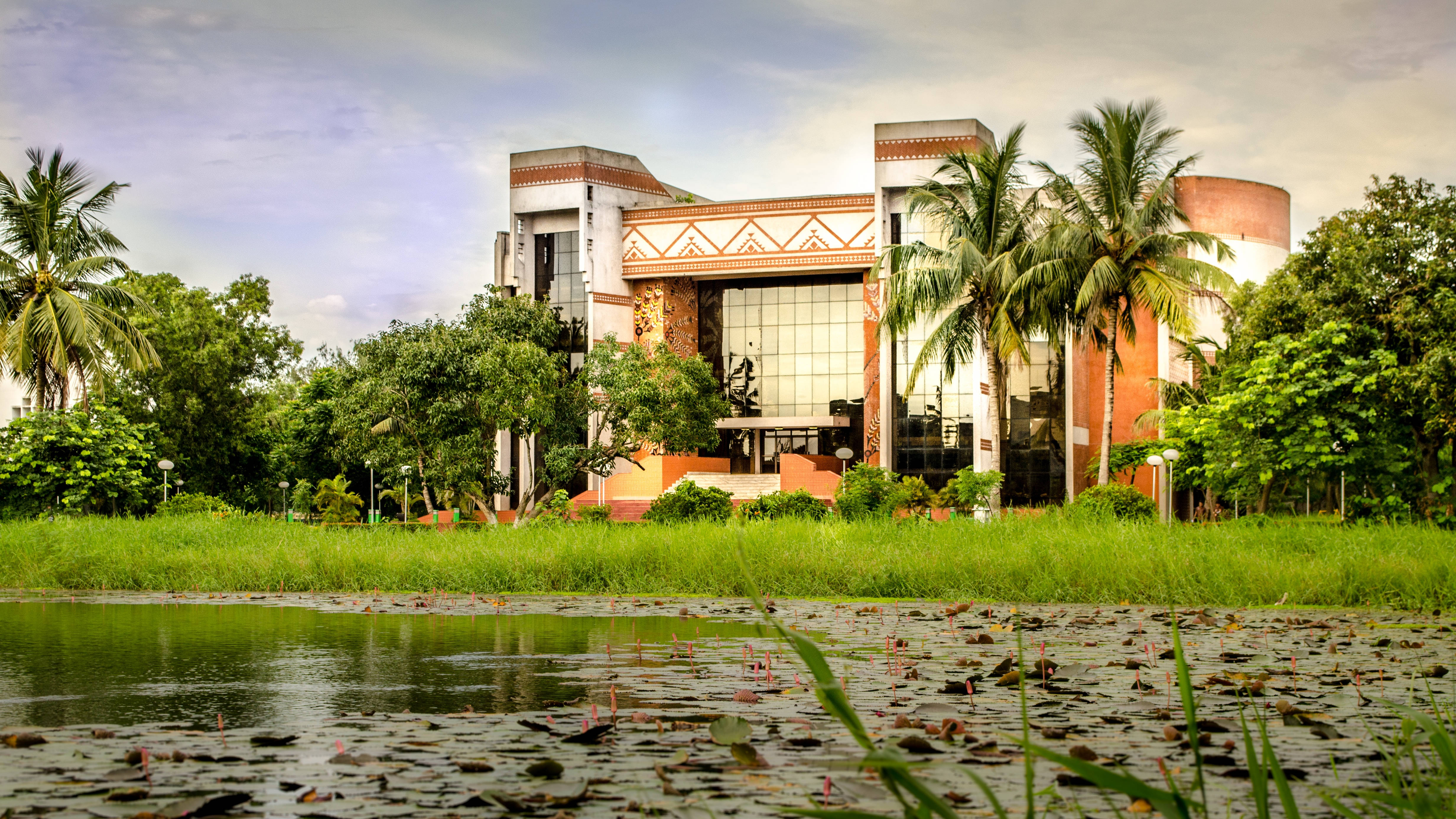Kolkata 桌布 壁紙 — 一座現代建築，帶有陶土色調和玻璃窗，矗立在茂密的綠草和點綴著睡蓮的池塘後面，天空柔和。